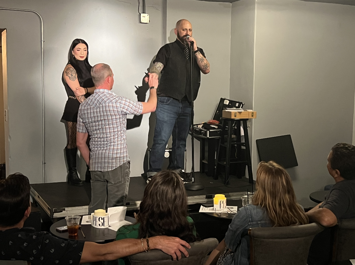 A man on stage with a microphone, wearing a black vest and jeans, holds hands with an elderly man in a plaid shirt. A woman with dark hair, dressed in black with visible tattoos, stands behind them. Several audience members sit at tables watching the
