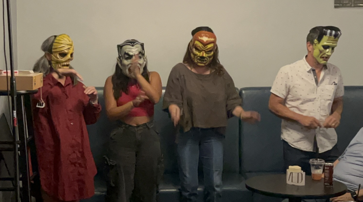 Four people wearing Halloween masks dancing in a room with a dark blue booth and a small round table with drinks.