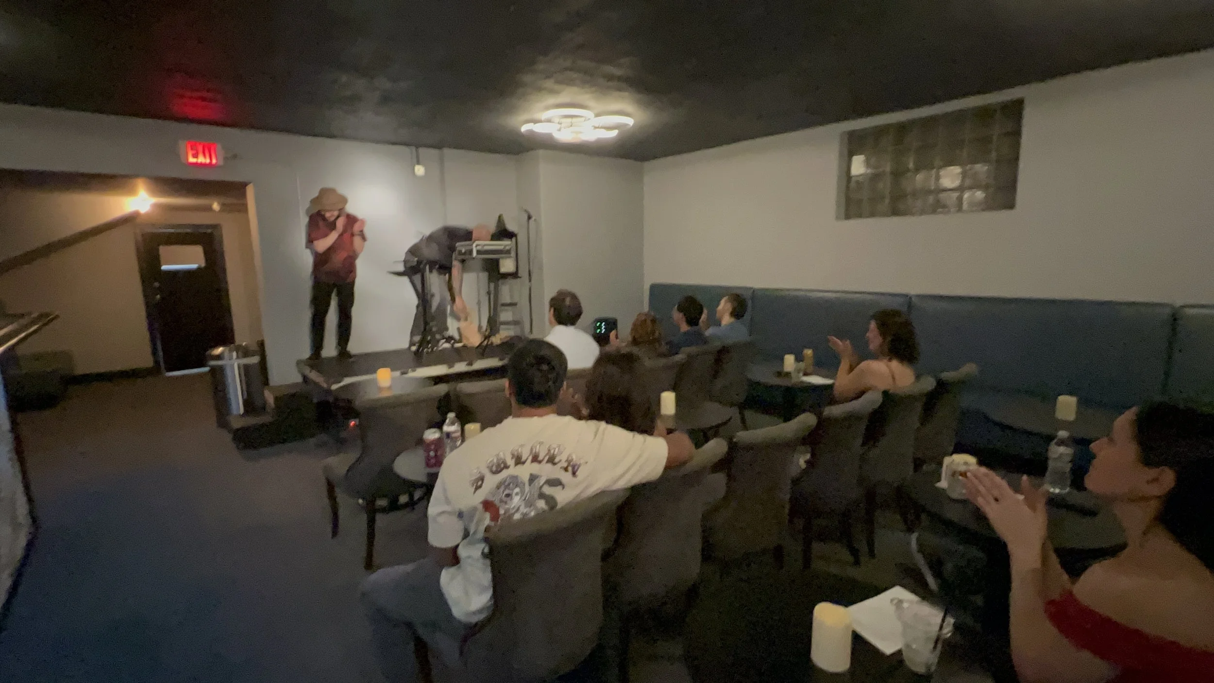 A group of people seated in a dimly lit lounge or bar, watching a person on stage performing or speaking, with some clapping and others looking on.