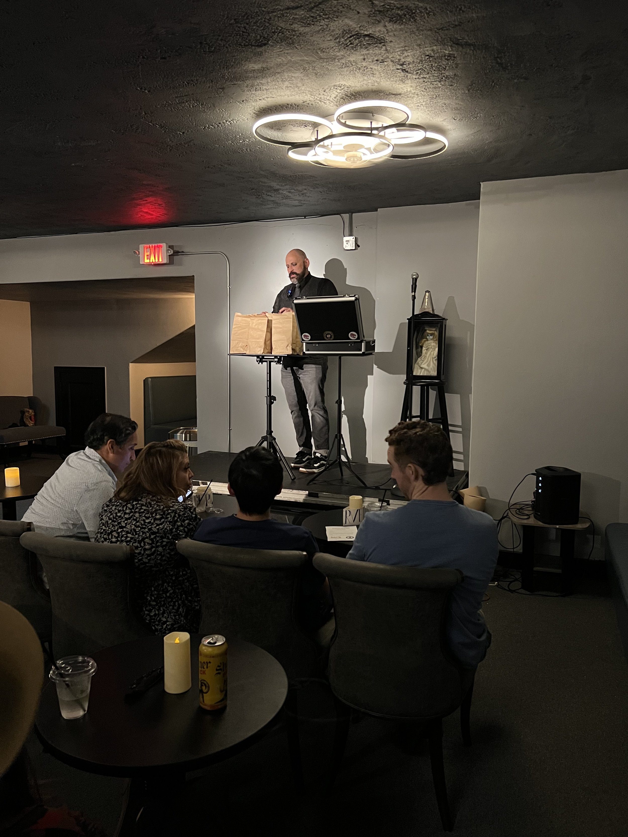 Man performing magic on stage at a restaurant or bar with three people sitting at a table watching.