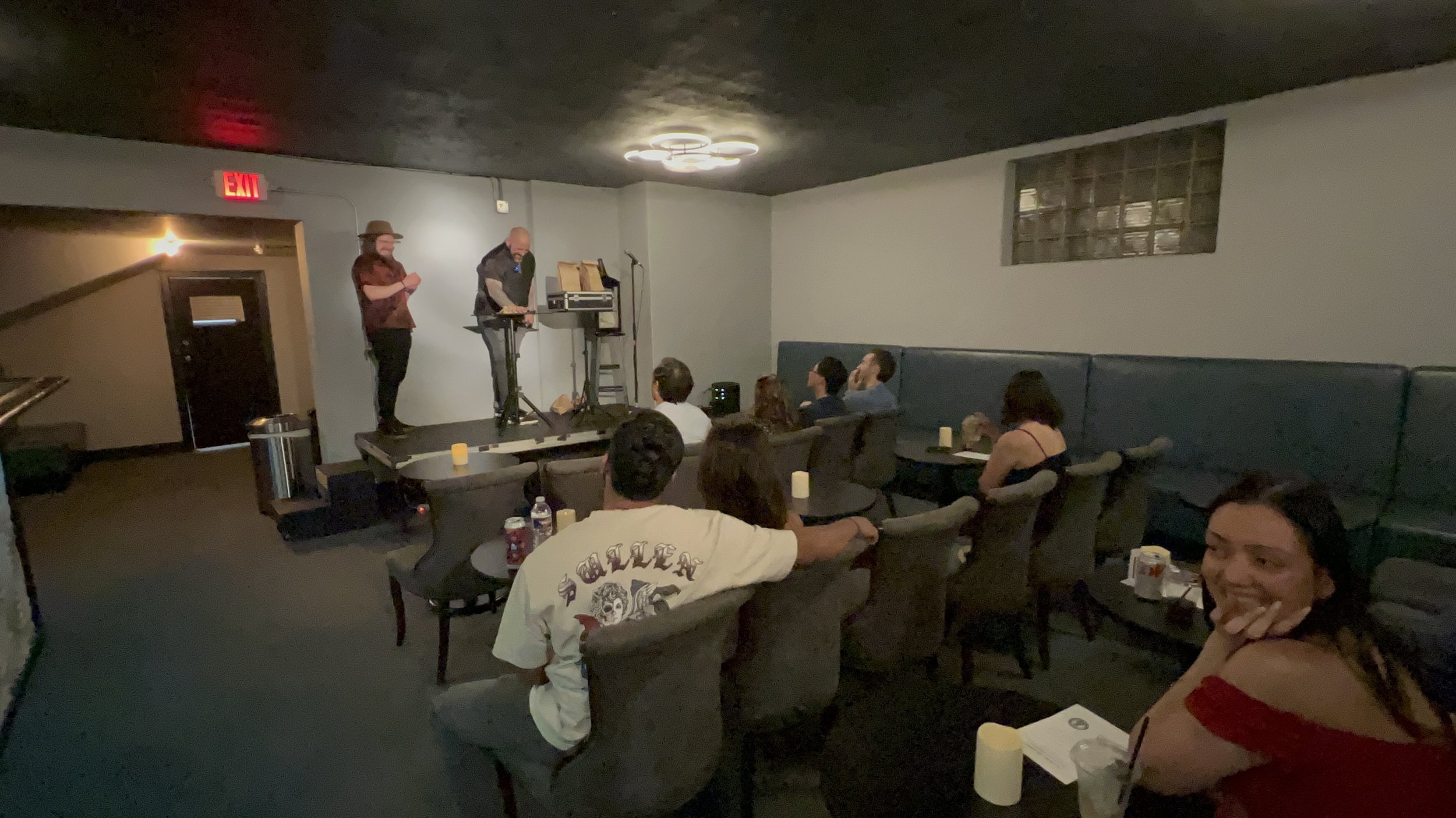 People watching a comedy performance or a presentation in a dimly lit room with a small stage, where two individuals are on stage with equipment, and audience members are seated on chairs and couches.