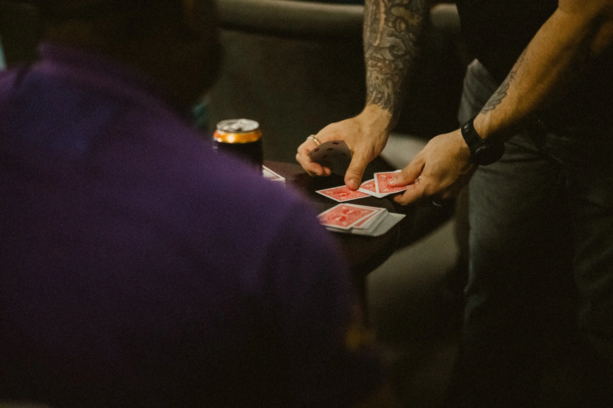 Two people playing cards at a table, one with tattoos on their arms, a deck of cards, and a soda can.