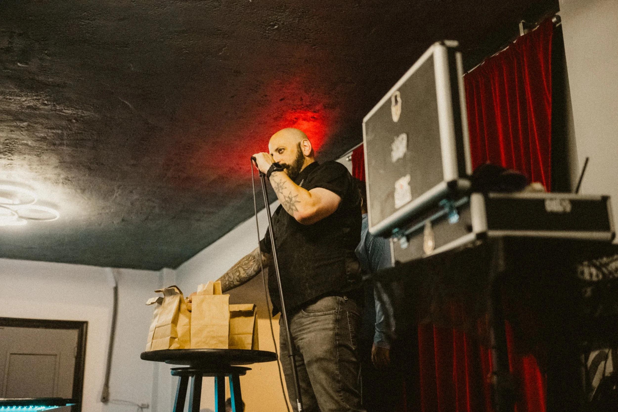 A magician with a shaved head and beard, holding a microphone, standing on a stage with a red curtain backdrop, next to a small table with paper bags.