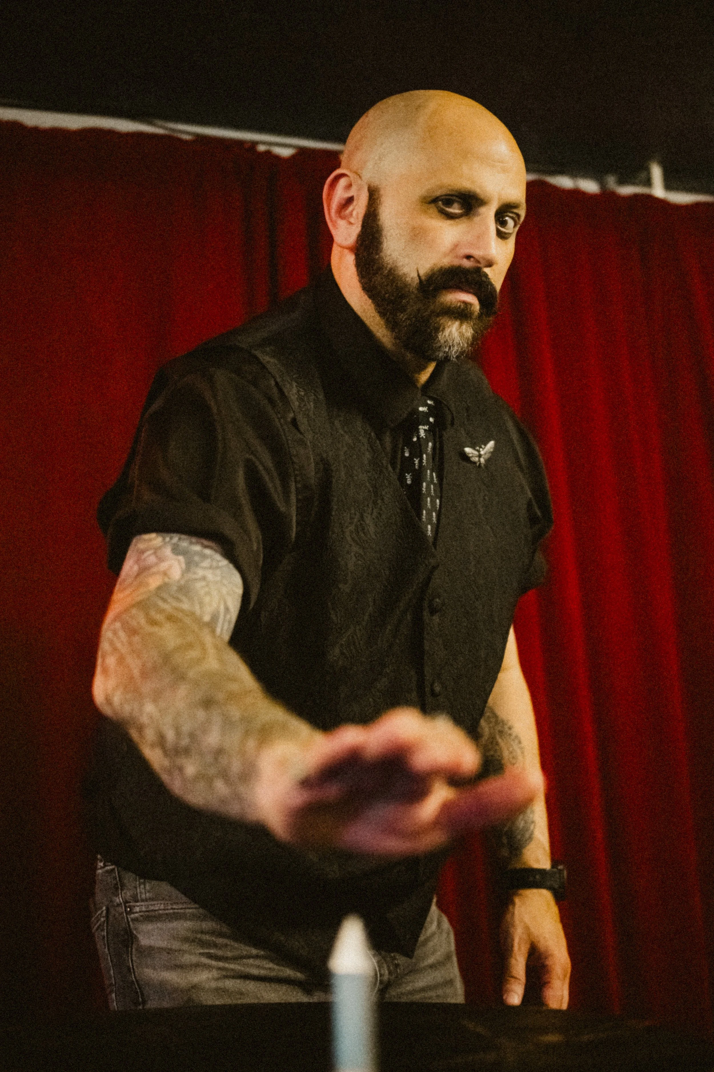 A magician with a bald head, beard, and tattoos wearing black clothing, reaching out towards a steel spike in front of a red curtain.
