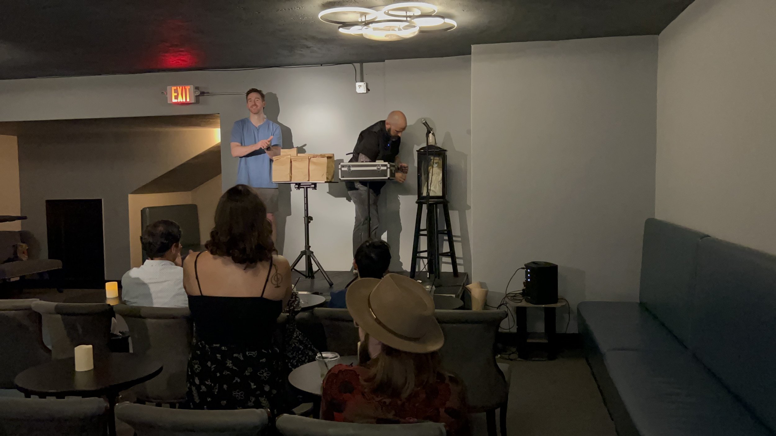 Two performers on stage, one with a microphone and others with a table and sound equipment, entertaining an audience in a dimly lit room.