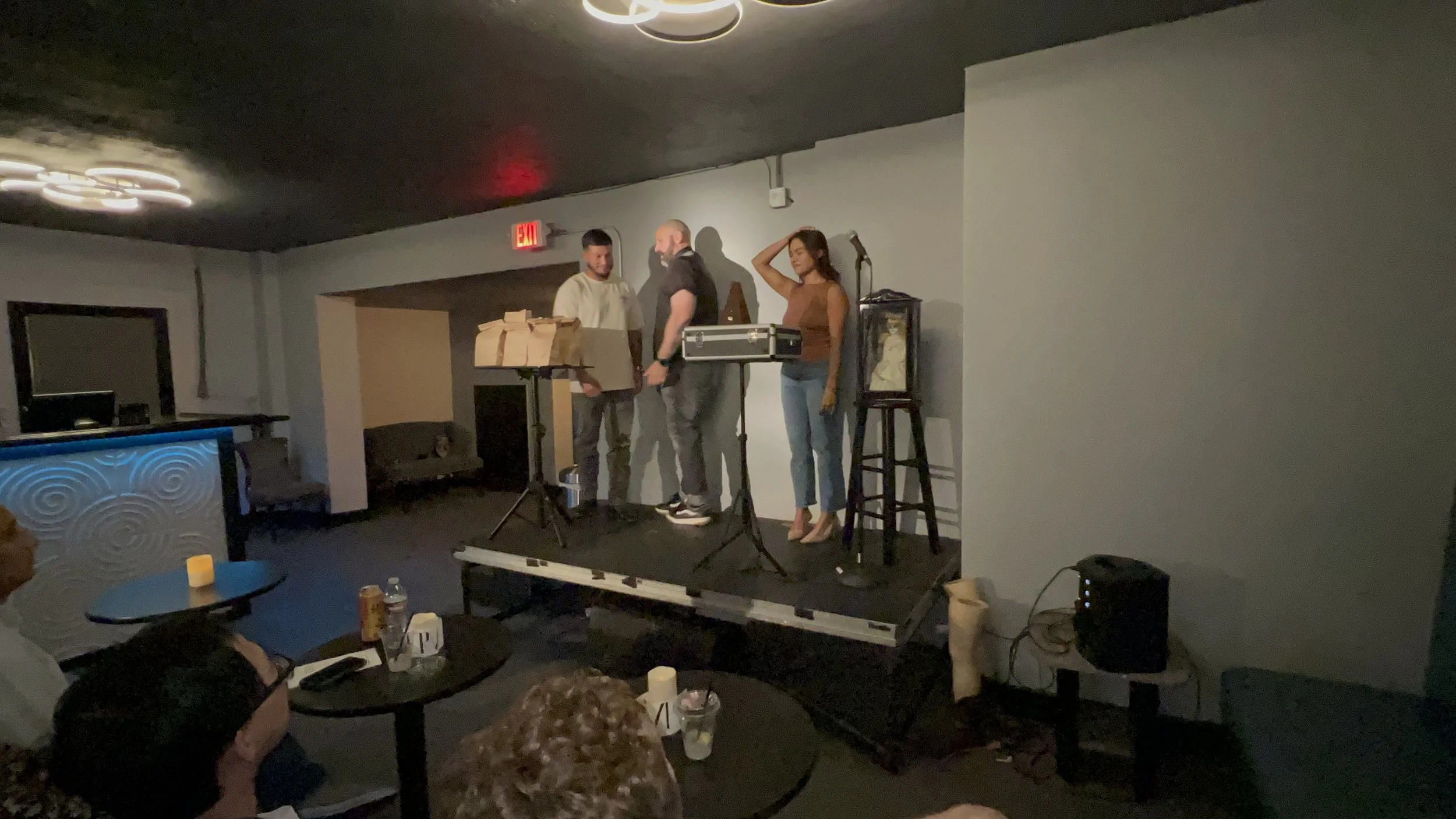 Three people on a small stage, two men and one woman, appear to be performing or rehearsing. One man is holding a microphone, another man looks at him, and the woman stands with one hand on her head. There are music stands and a microphone on stage. 