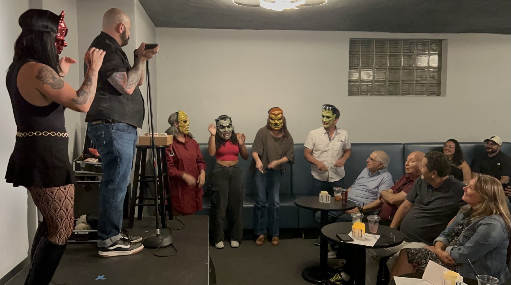 Group of people in a room attending a comedy show or performance. Three performers on stage wear masks resembling horror or fantasy characters, while a woman on stage with a red top and black pants talks or gestures. The audience is seated at tables,
