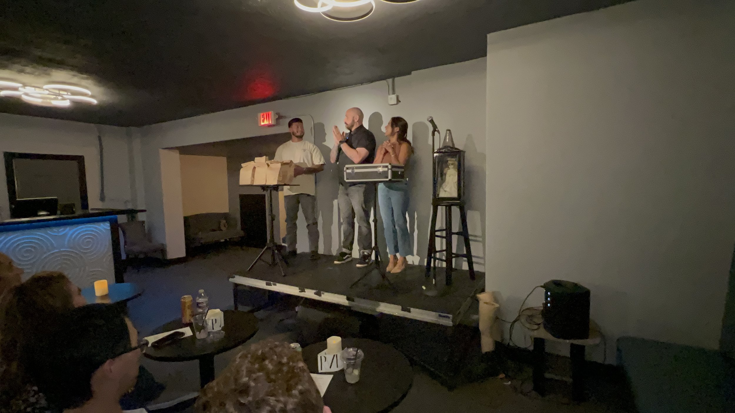 A group of three people standing on a small stage, with two men and one woman in conversation. The stage has microphones, a treatment case, and a stool. Audience members are seated at tables in front, watching. The room has dim lighting and modern de