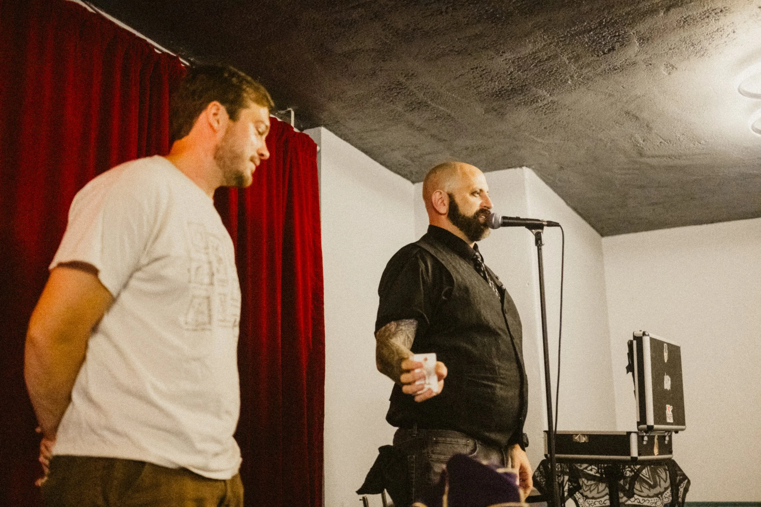 Two men on stage, one with a microphone and tattooed arm, the other standing with hands behind back, in front of a red curtain, with audio equipment on side.