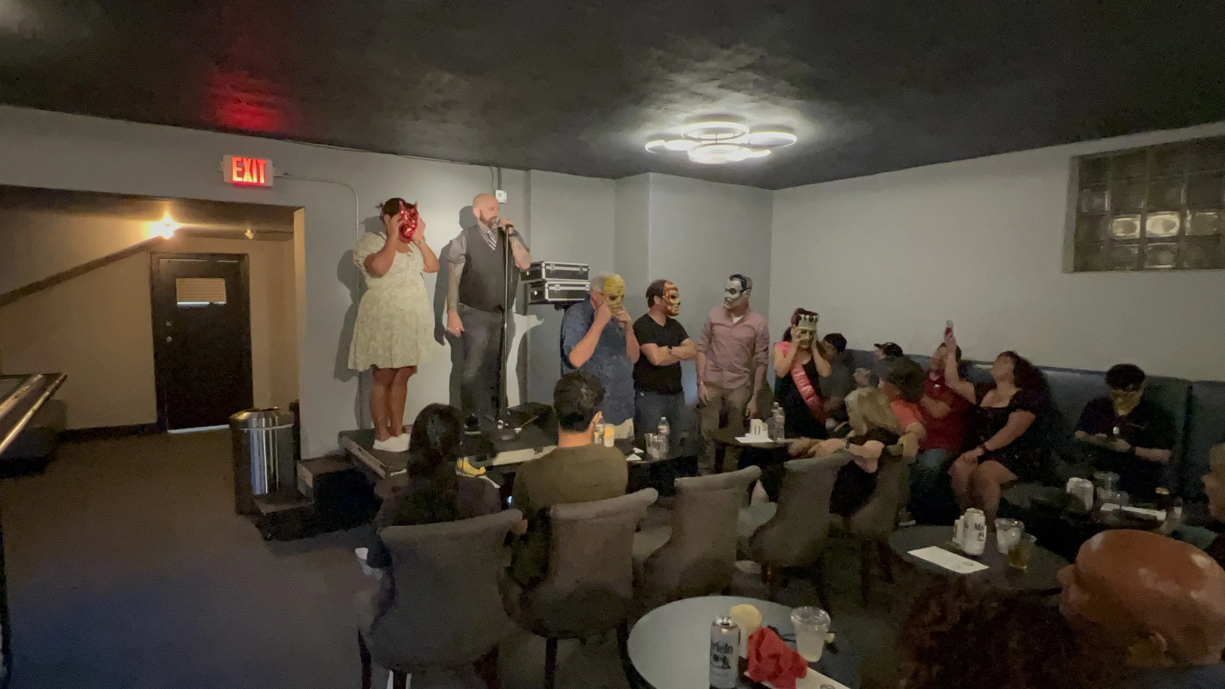 People wearing masks and costumes in a dimly lit room during a gathering or performance, with some standing on a raised platform and others seated at tables.