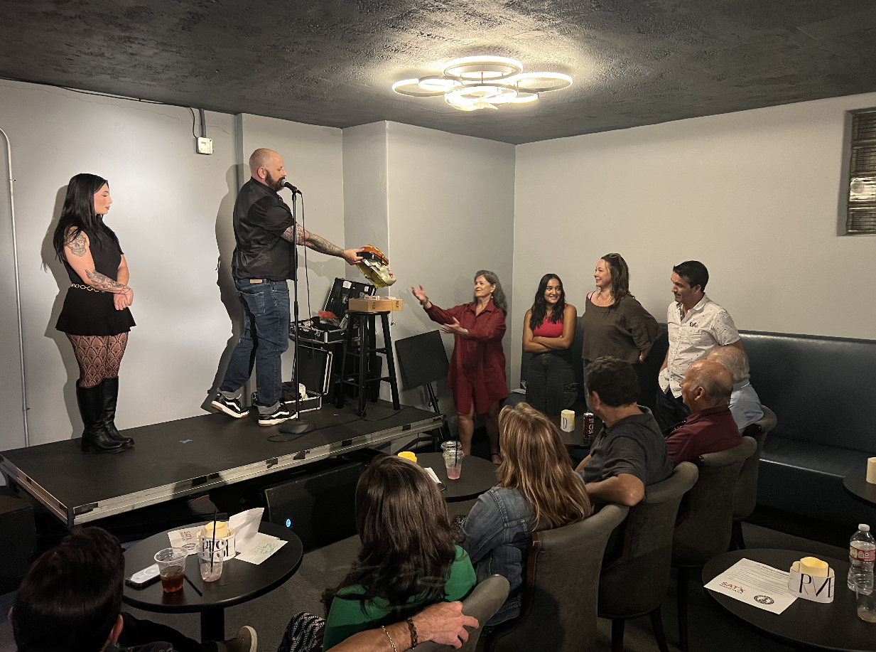 Group of people on stage at an event, with one person holding a hamburger prop, and others engaging, while an audience watches.