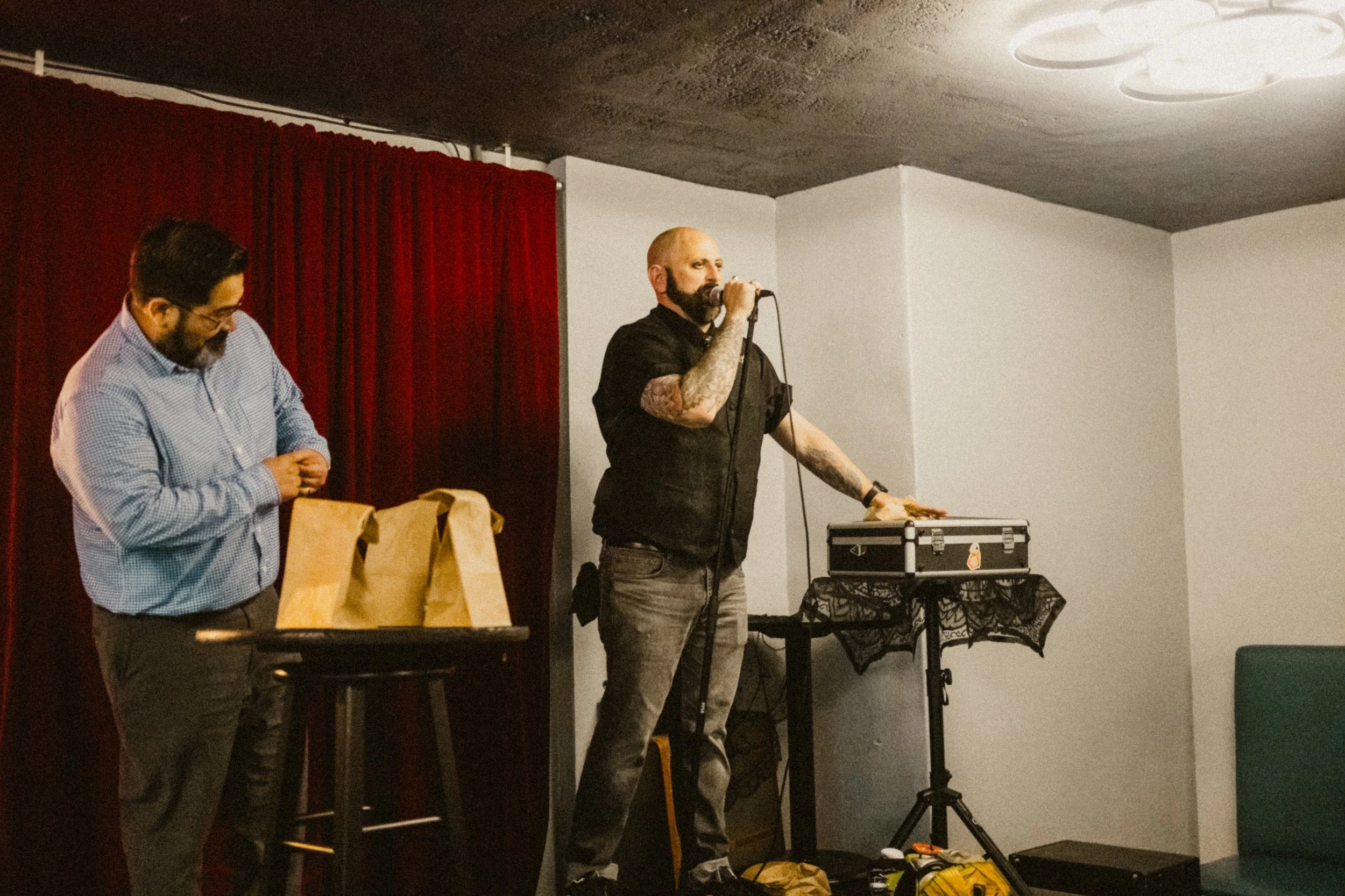 A man with tattoos and a black shirt is standing on a small stage, holding a microphone and resting his hand on a DJ setup. A second man in a checked shirt and glasses is standing nearby with a paper bag, looking down.