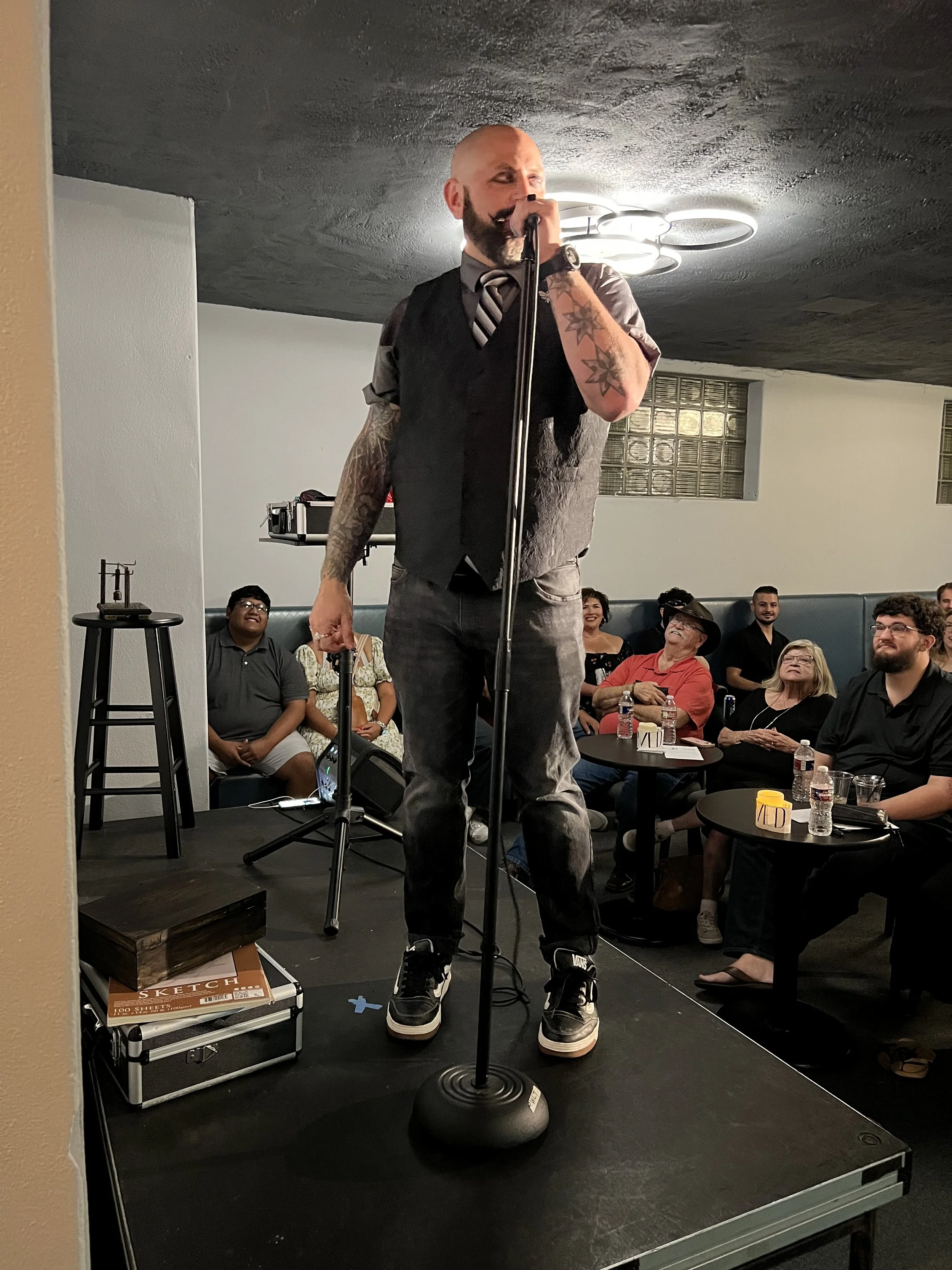 A man with tattoos and a beard stands on a small stage, holding a microphone and performing in front of an audience seated at small tables, with some people smiling and laughing.