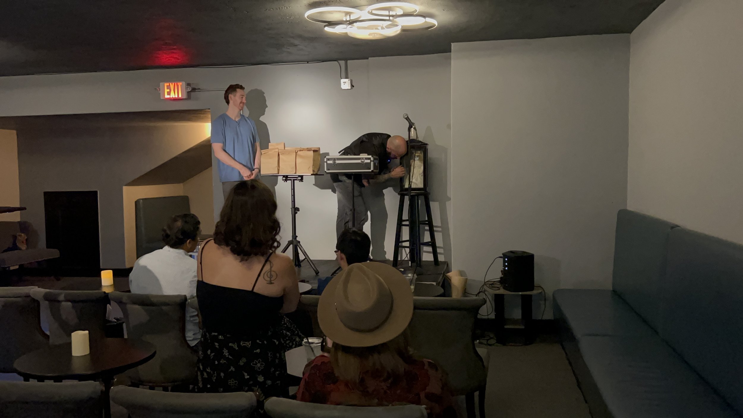A live performance in a small venue with a man playing a musical instrument on a stage, a man standing next to him, and an audience watching seated. The room has minimalist decor with a black ceiling, modern lighting fixture, and a seating area with 