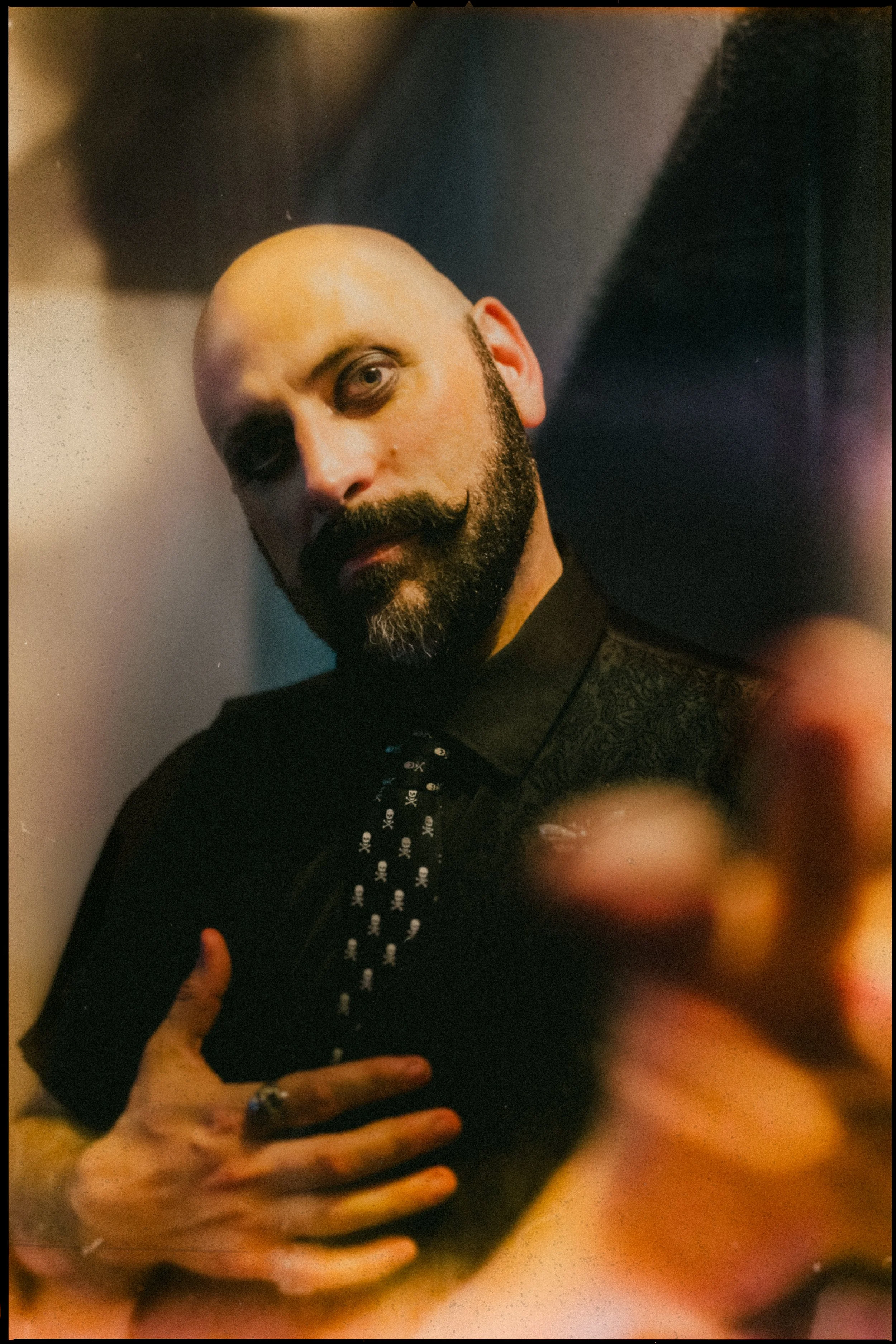 A bald magician with a beard and mustache, wearing a dark shirt and tie, looking at the camera with a slightly tilted head, with the person's hand in the foreground and an out-of-focus background.