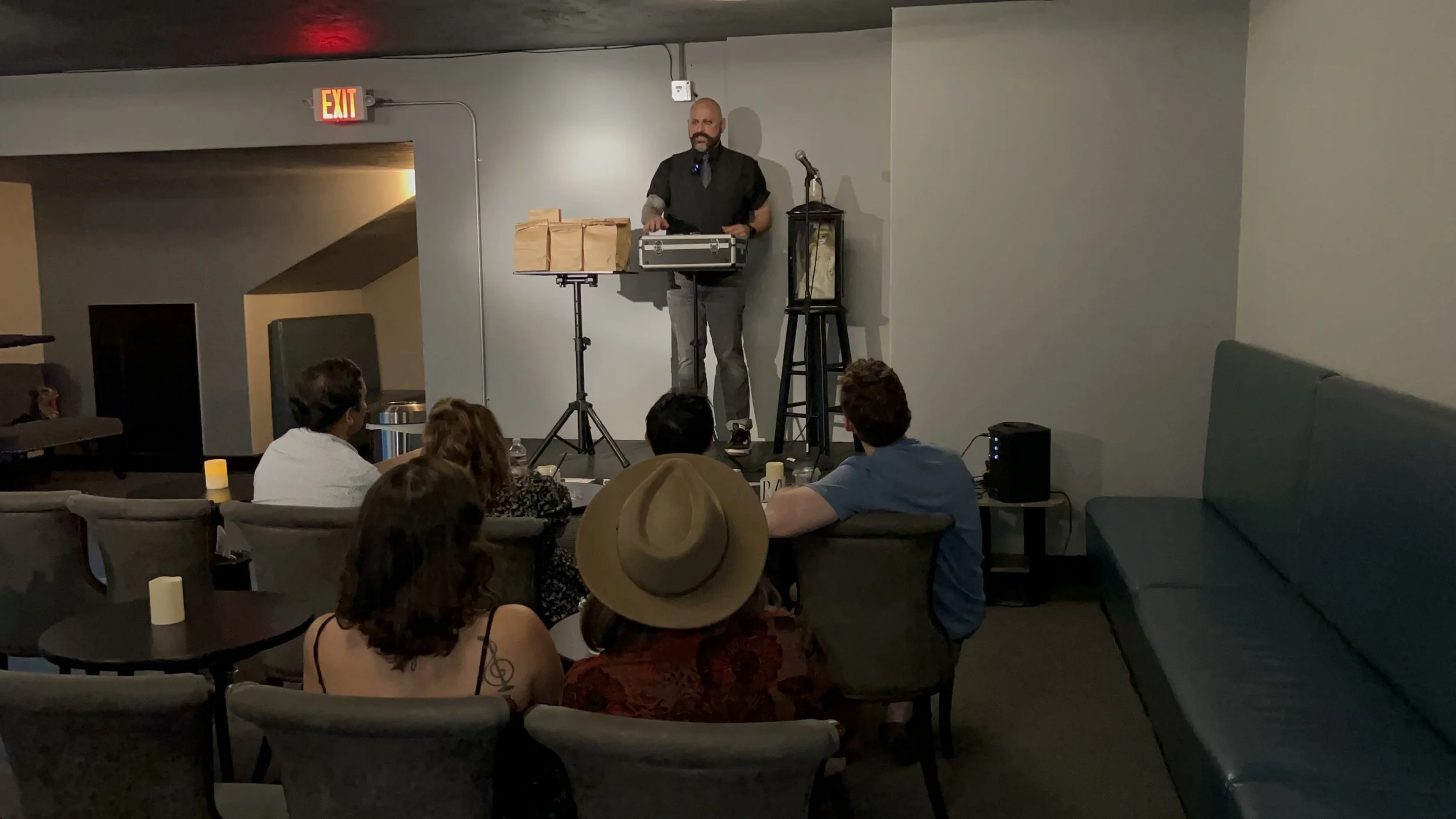 A man standing on a small stage with a microphone and music equipment, performing or speaking for an audience of five people seated in a dimly lit room, with a large couch on the right and various decorations and lighting around.