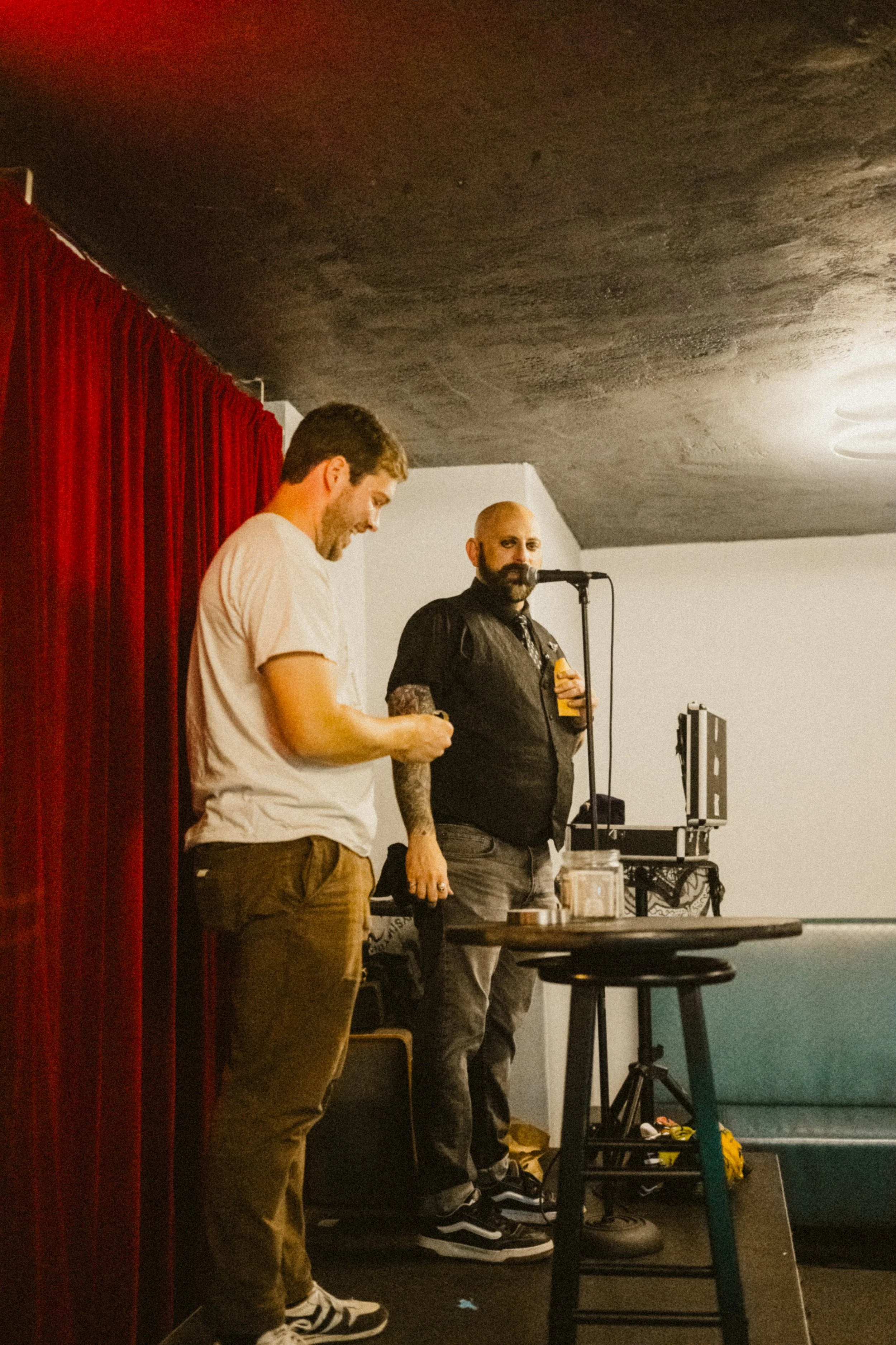Two men standing on a small stage with a red curtain and a black ceiling. One man is looking at his phone while the other is holding a drink and looking at the camera. There is a microphone, a small table, and some equipment behind them.