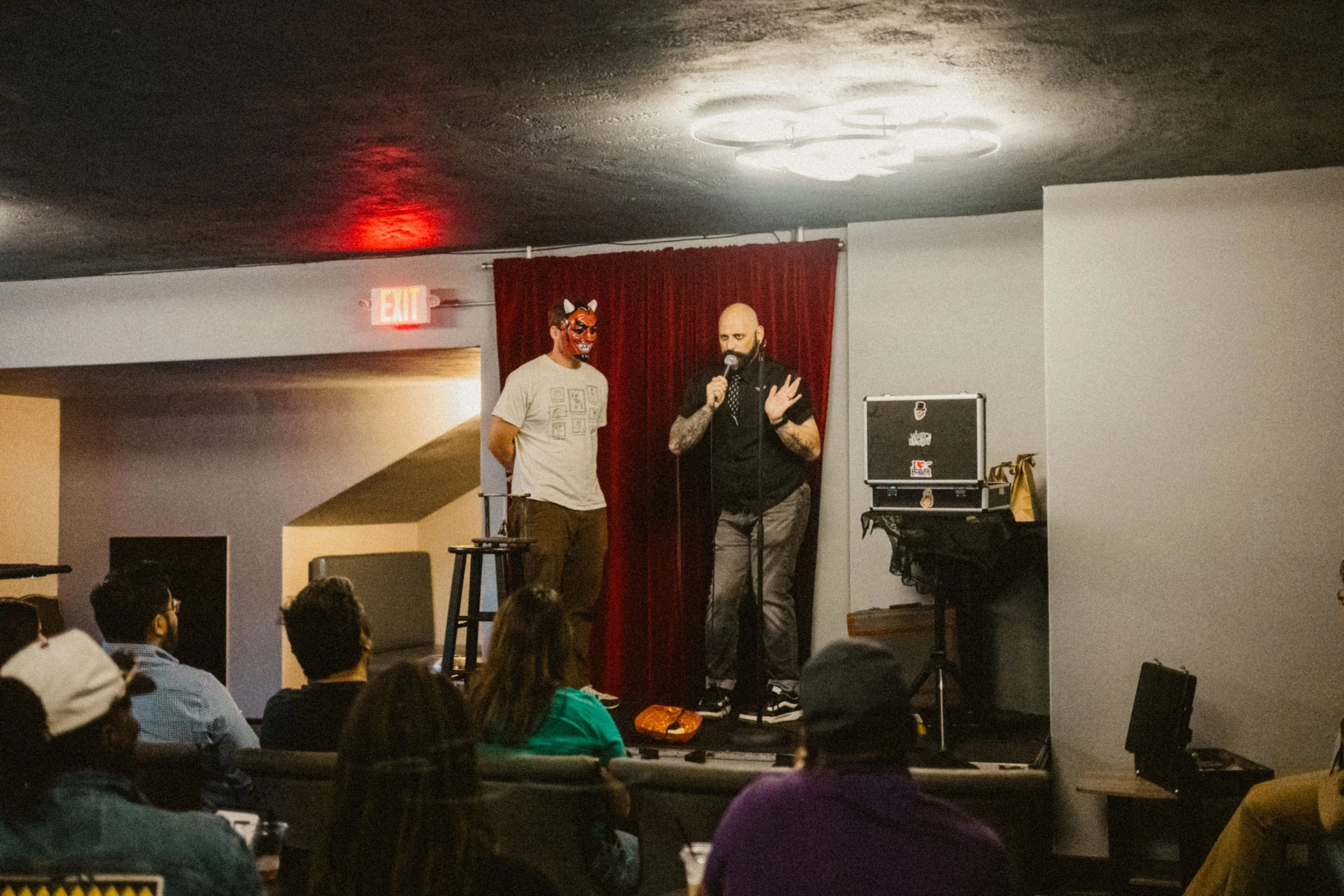 A comedy magic show on stage with audience member wearing a mask and casual clothes, and the magician is bald with a beard, tattoos, black shirt, and gray pants. Audience members are seated watching the performance.