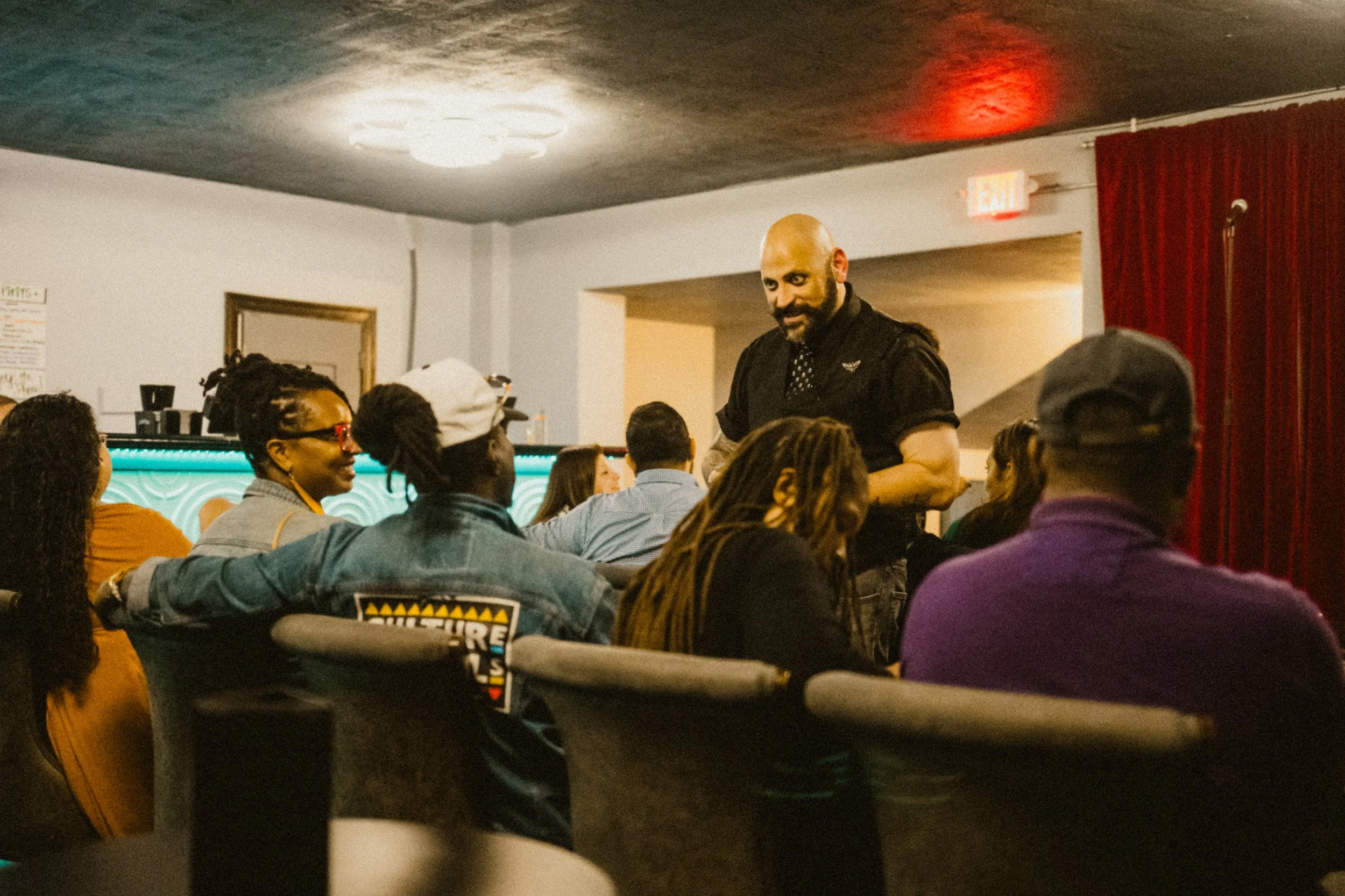 A magician with a bald head and beard standing and talking to a group of seated people in a dimly lit room, with a red curtain and exit signs in the background.