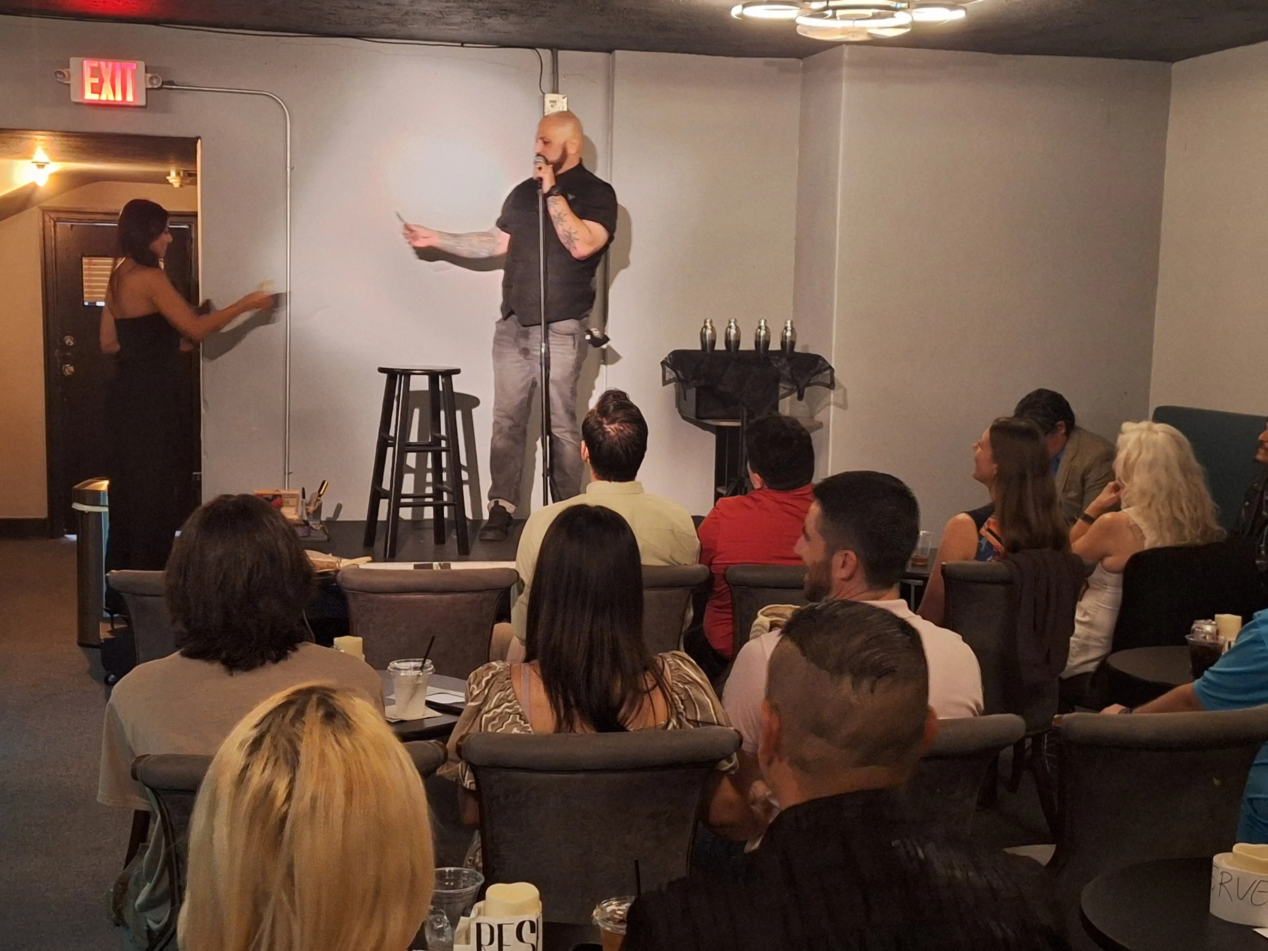 A man with tattoos on his arms is speaking into a microphone on a small stage in front of an audience. A woman is standing near a door to the left, holding a small paper or card. The audience is seated at tables, watching the performance or speech.