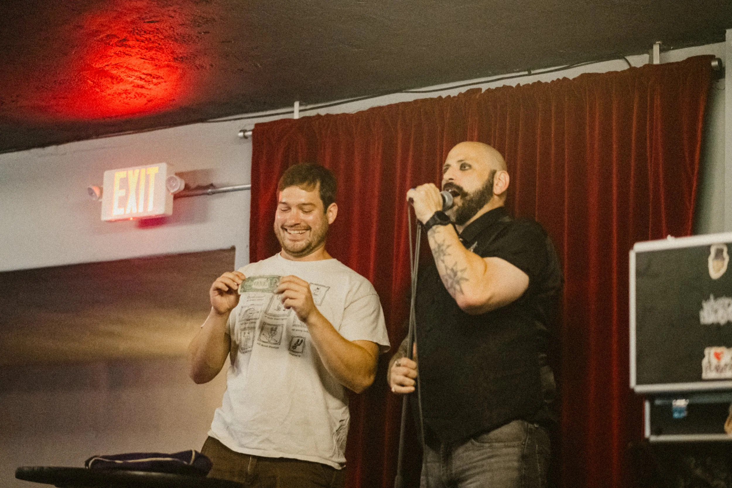 Two men on stage, one holding money, are smiling and laughing, with a red curtain and an exit sign in the background.