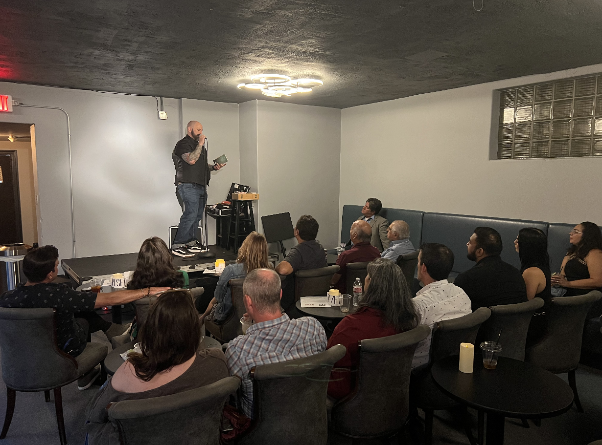 Man on stage reading from a book to an audience in a dimly lit room with gray walls and modern ceiling lights.
