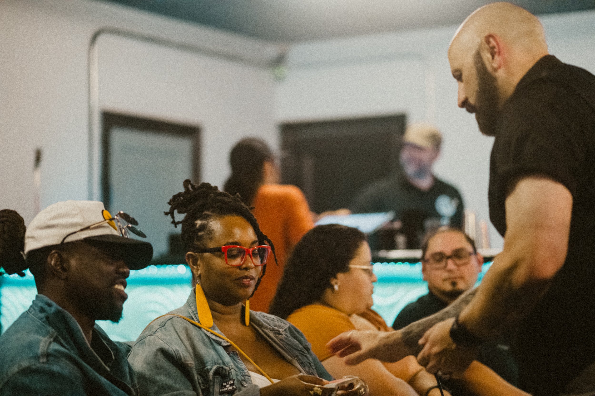 A group of people seated indoors, engaging in conversation. One man with a shaved head and beard is talking to a woman with glasses and bright red earrings, while others look on. The background shows a bar with people and a DJ.