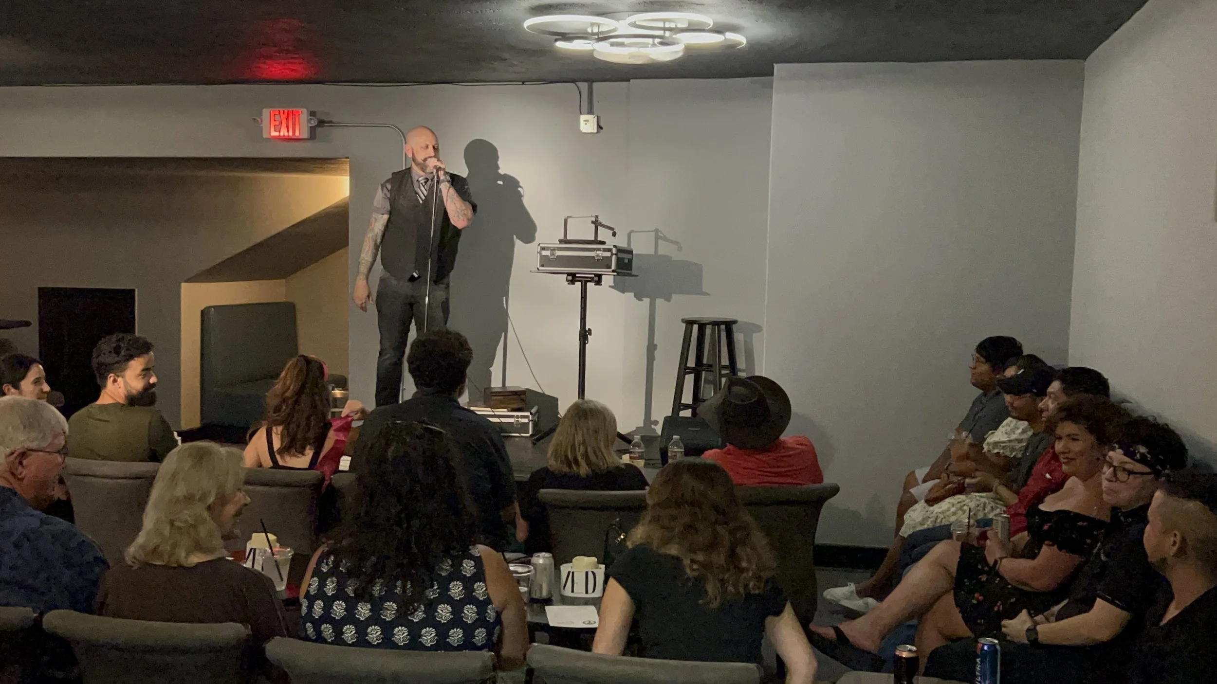 A man on a stage with a microphone singing in front of an audience at an indoor venue, with a small equipment stand and a stool on stage, audience members seated and watching.