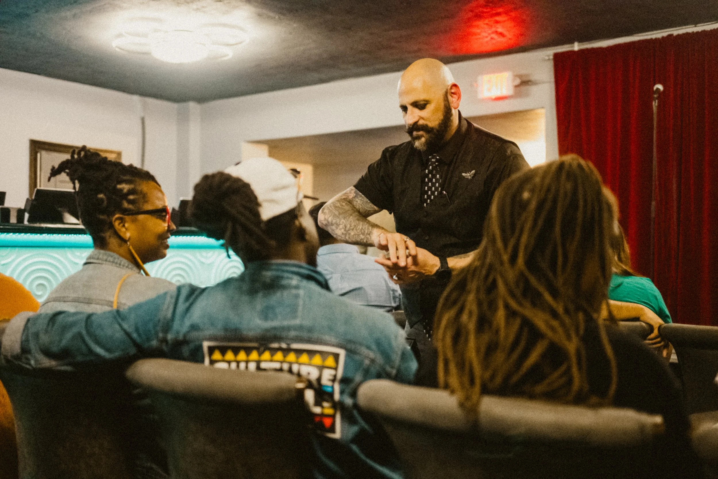Magician with a bald head, beard, and tattoos talking to a group of diverse people seated at a table in a casual setting, possibly a bar or cafe.