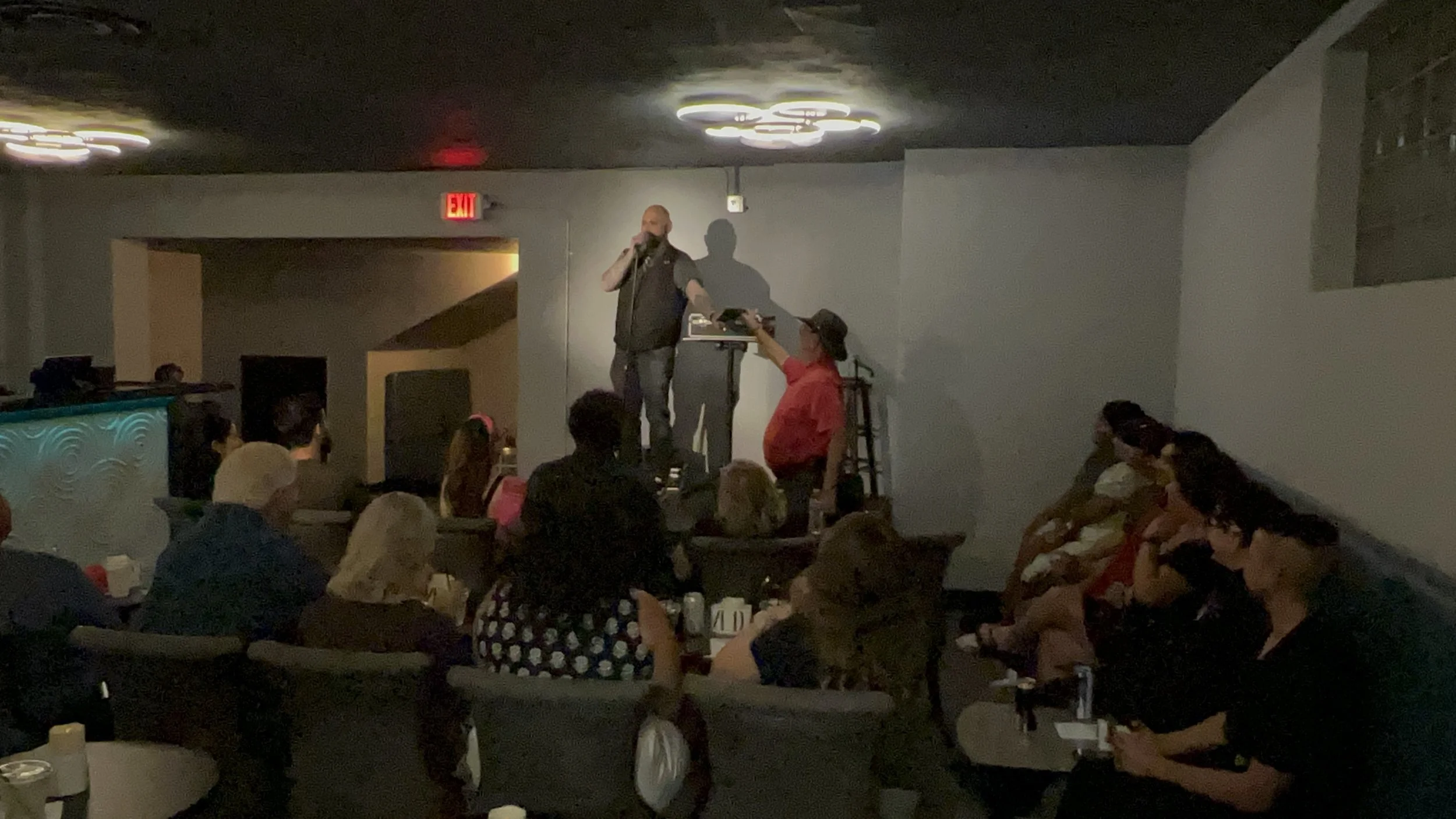 A man speaking into a microphone on a small stage with a DJ setup, indoors with dim lighting, audience sitting on couches and chairs watching.