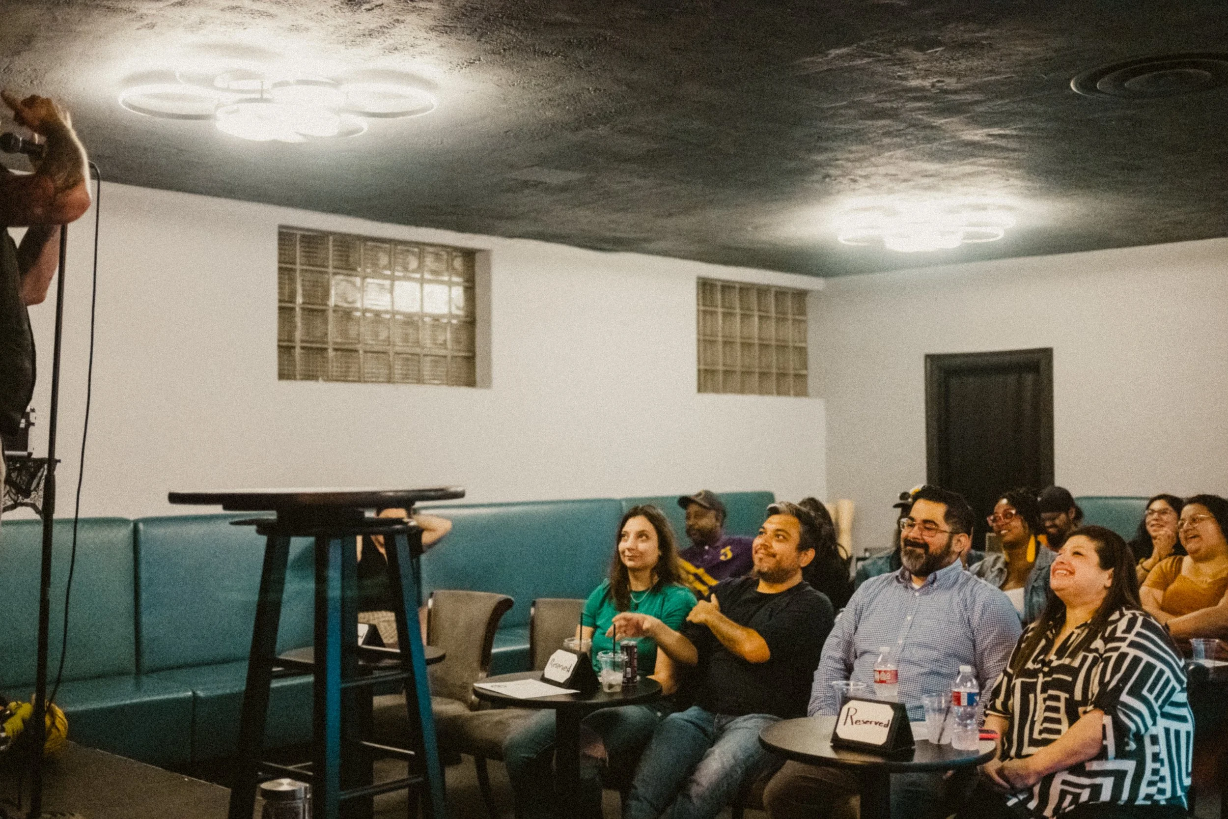Audience members seated in a room watching a magician on stage with a microphone. The room has a white wall, teal chairs, and two glass block windows.