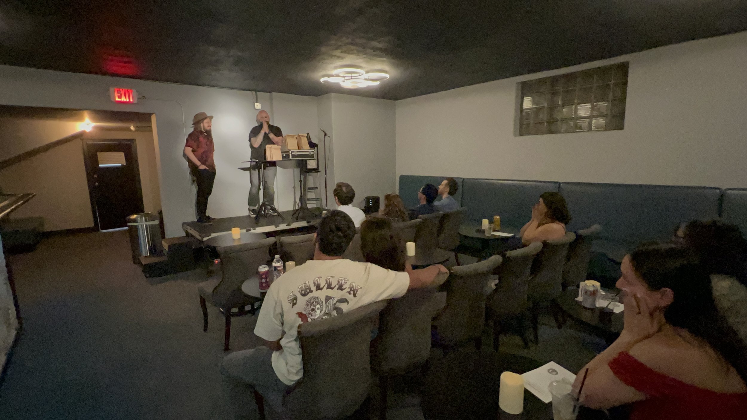 People watching a performer on a small stage in a dimly lit room with a black ceiling and blue seating.