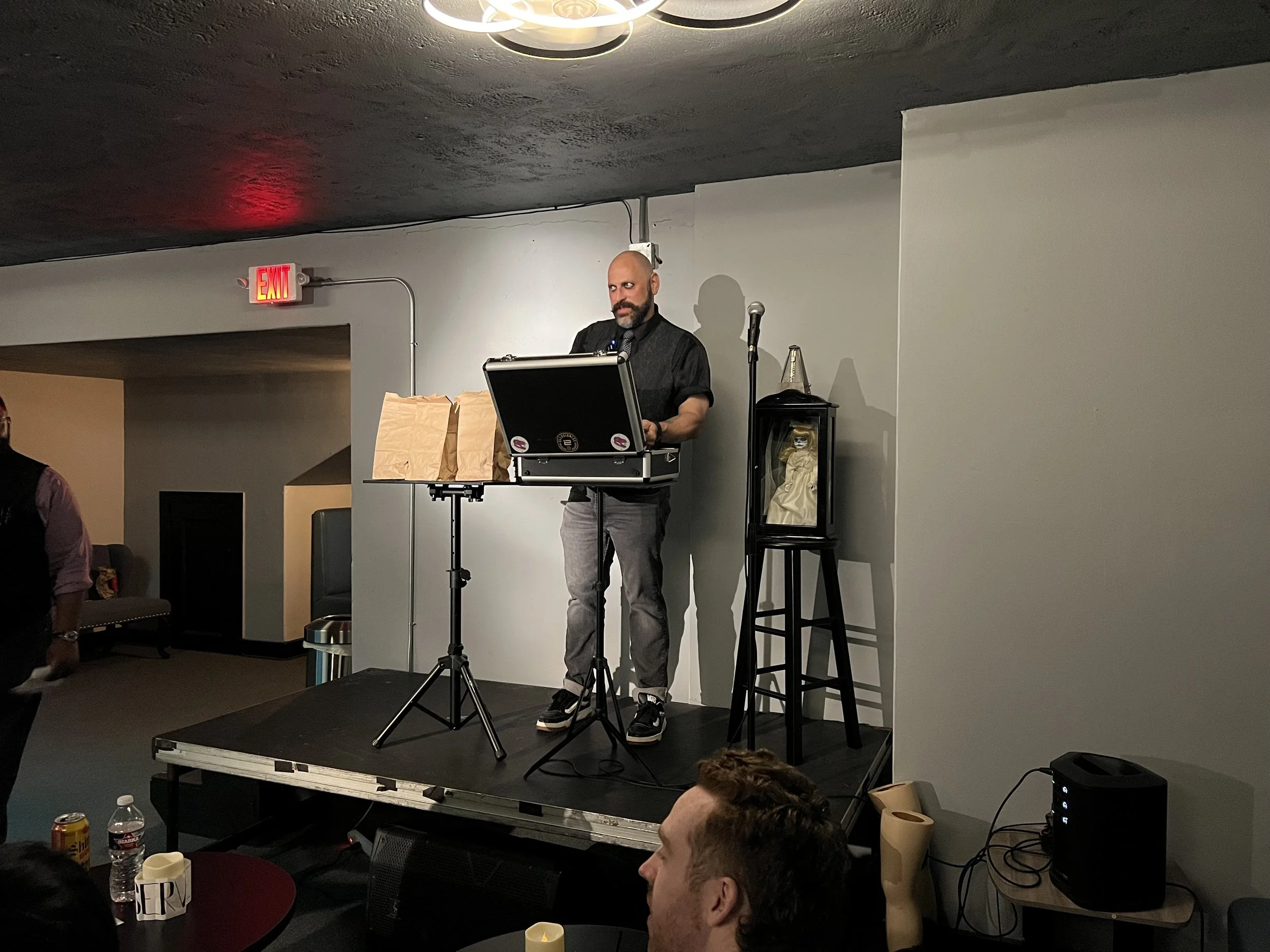 A man standing on a small stage with a microphone and a laptop, in a dimly lit room. There are paper bags on the stage, a small statue or decorative figure inside a glass case on a stand, and a speaker on the floor. Audience members are visible, with