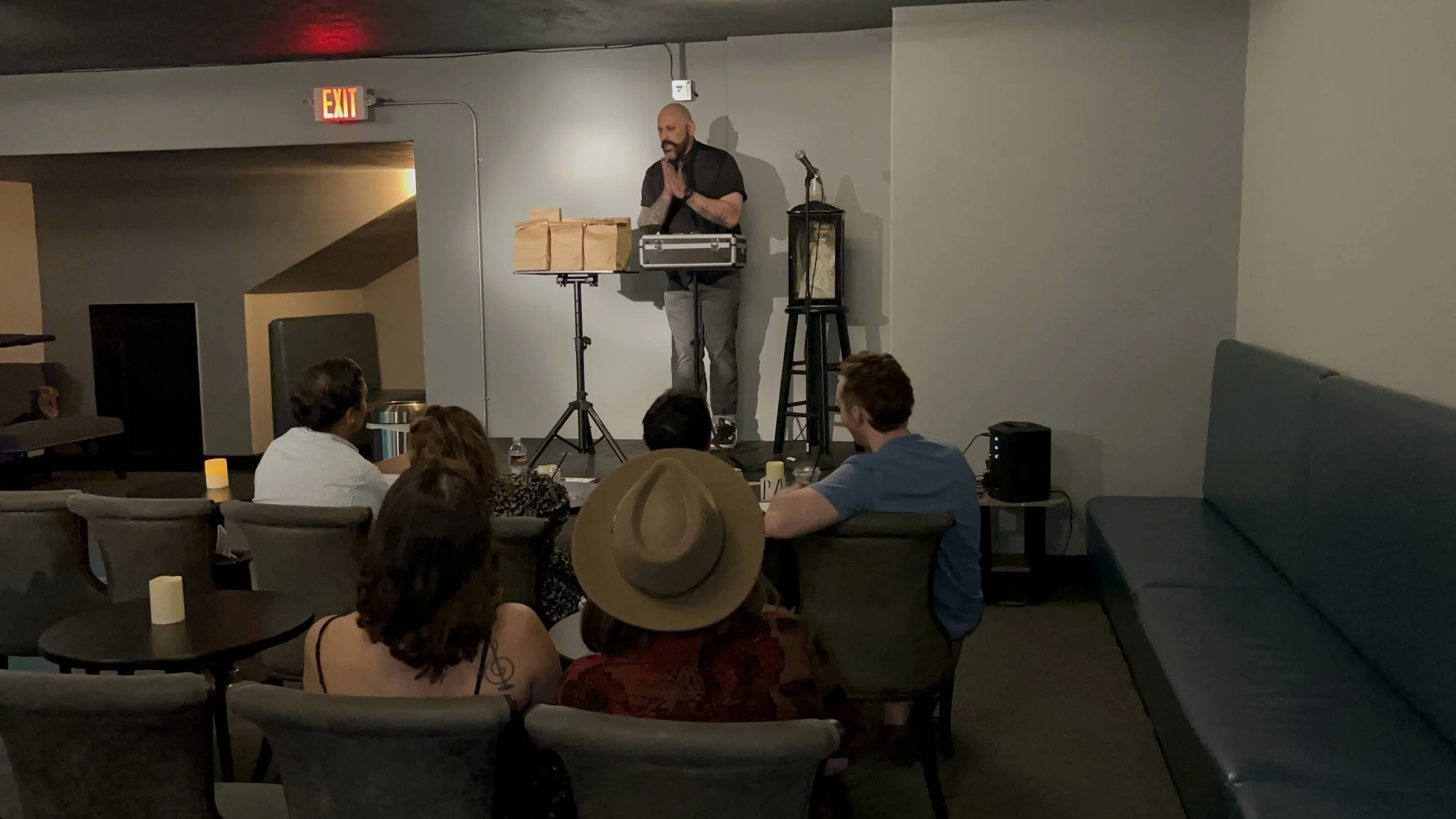 A man with a bald head, beard, and tattoos on his arms is performing a comedy stand-up act on a small stage in front of an audience seated at tables. The audience includes a person wearing a large tan hat. There are several small candles on the table