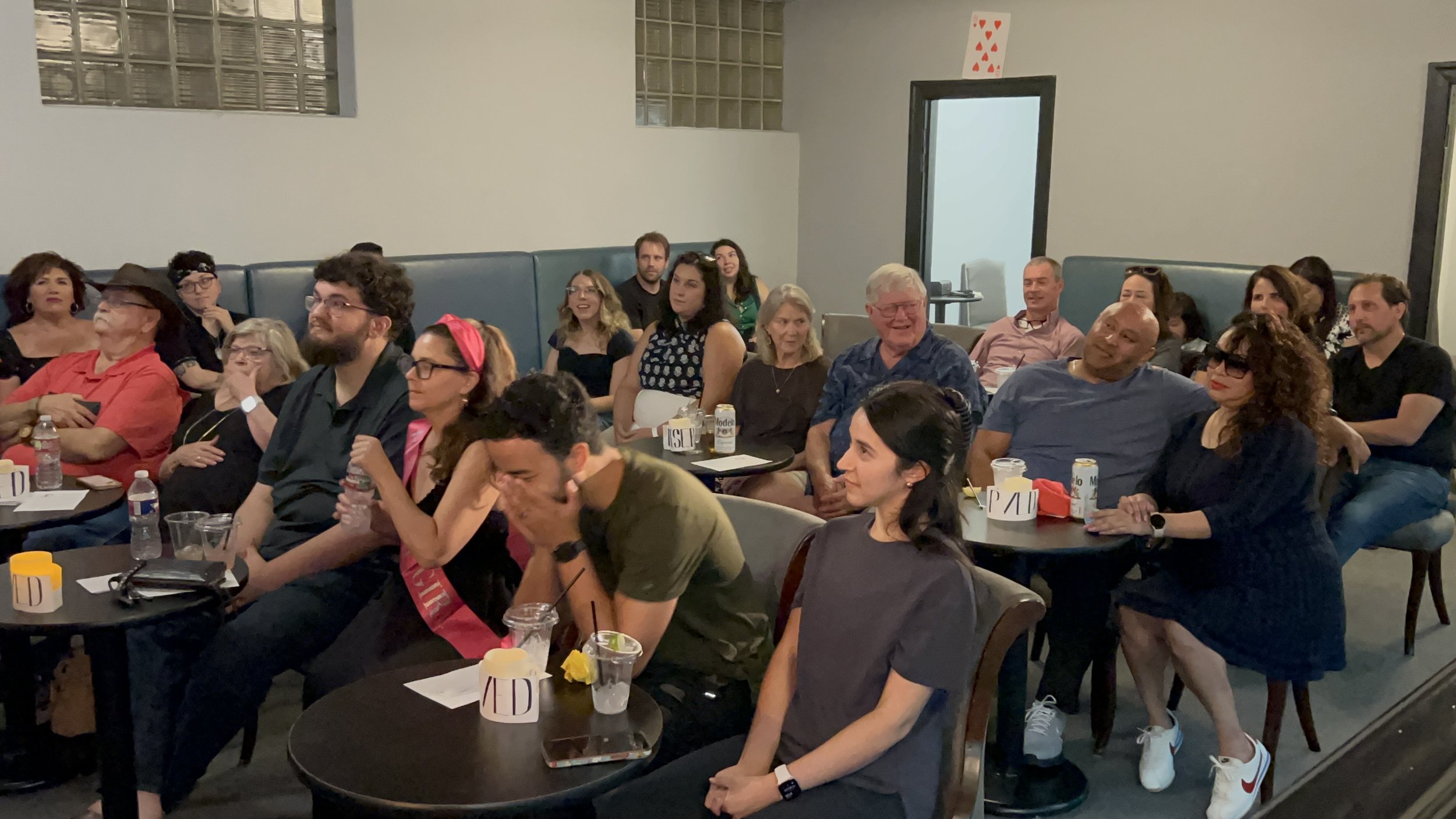 A group of people sitting in a conference room, laughing and enjoying a presentation or performance. They are at round tables with drinks, papers, and small signs, with a mix of men and women of various ages.