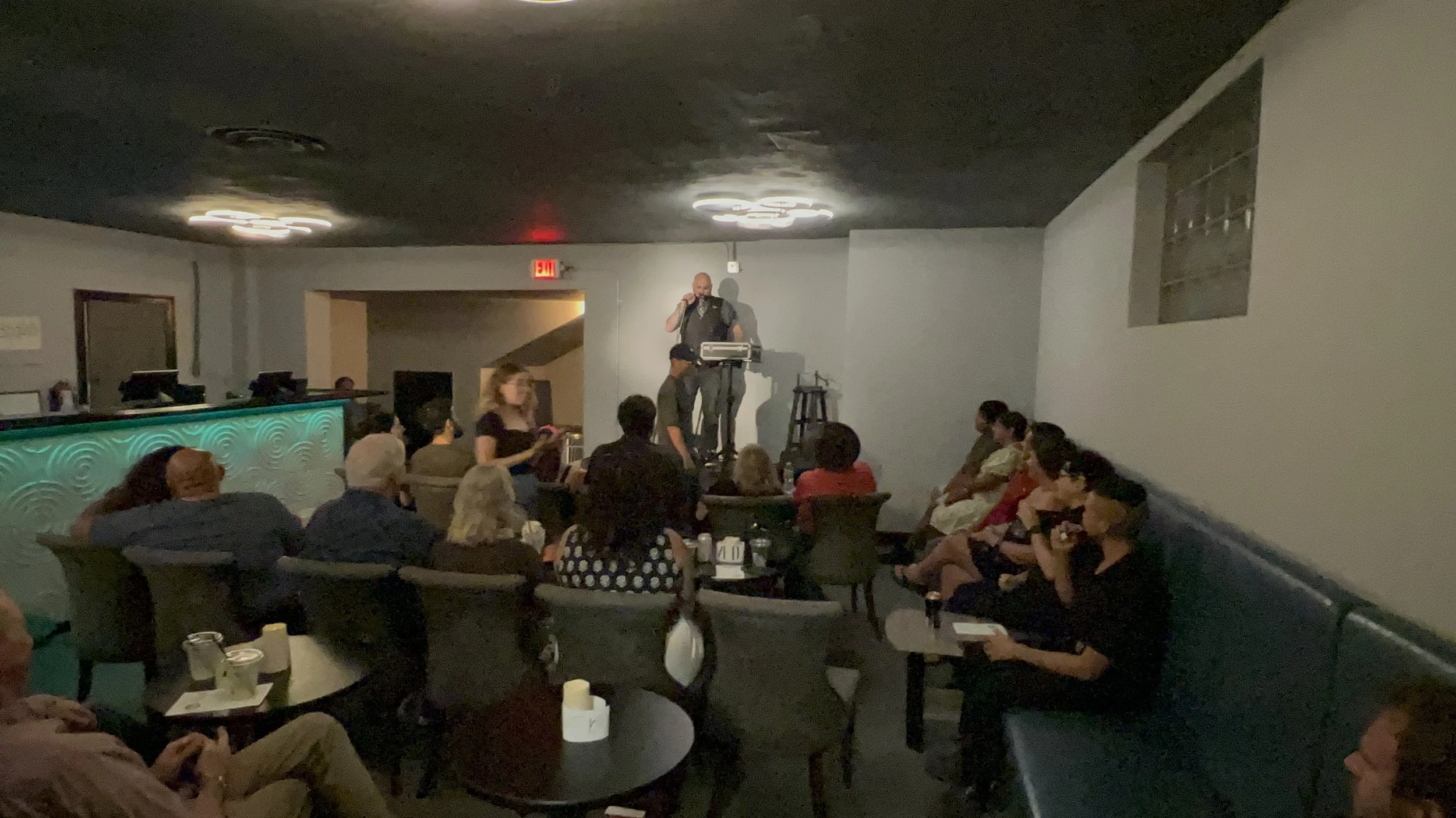 People sitting on chairs and couches in a dimly lit room, listening to a man performing or speaking on a stage with a microphone and stand.