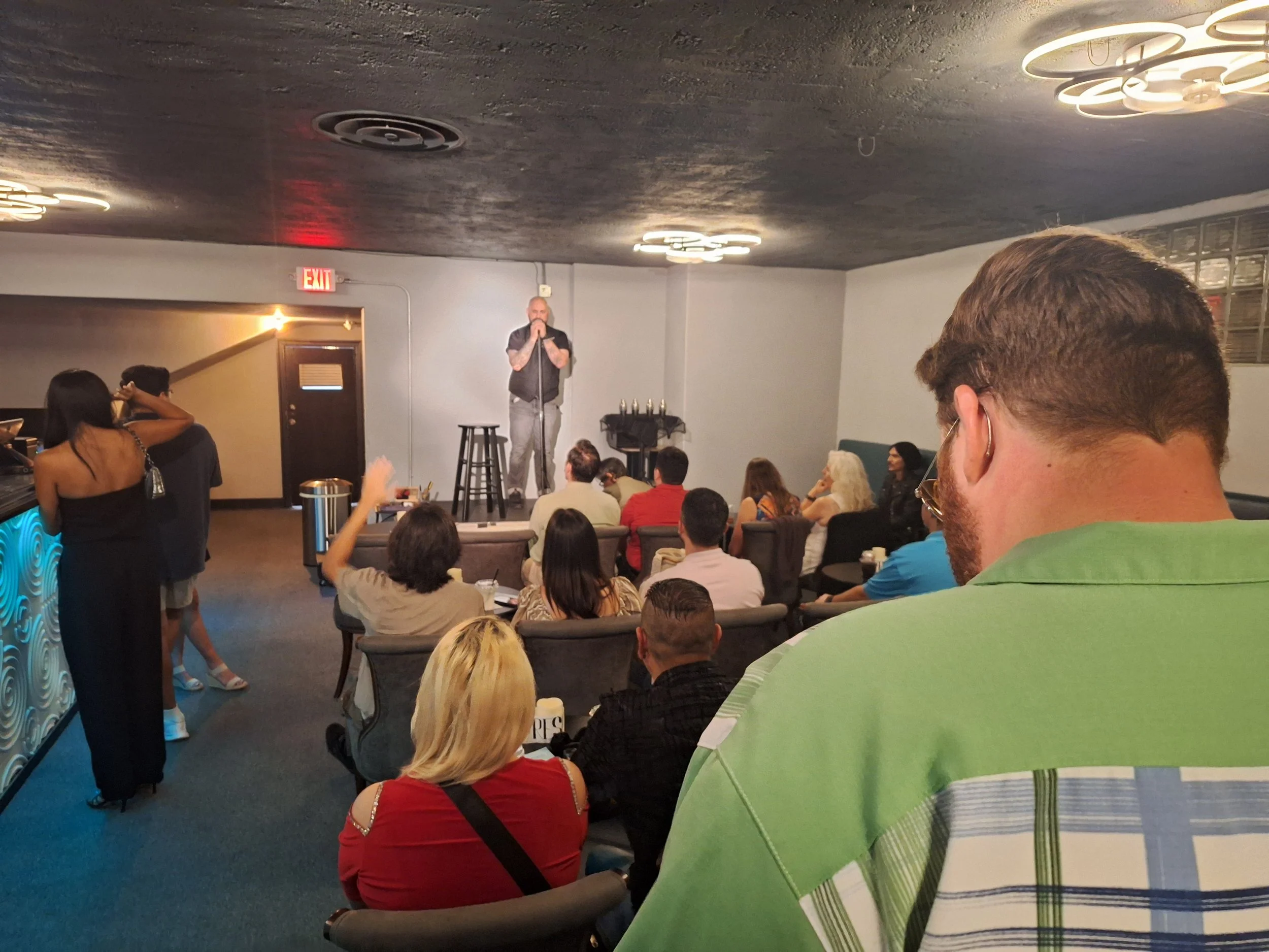 A comedy show in progress at a small venue with an audience seated on sofas and chairs, a male comedian on stage with a microphone, and a bar area with a woman and man near the drinks.