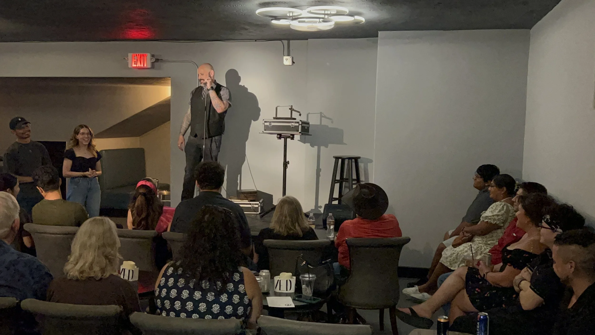 A comedian performs on stage in front of an audience at a comedy club, with some people standing on stage, while others sit and watch, and some audience members holding drinks.