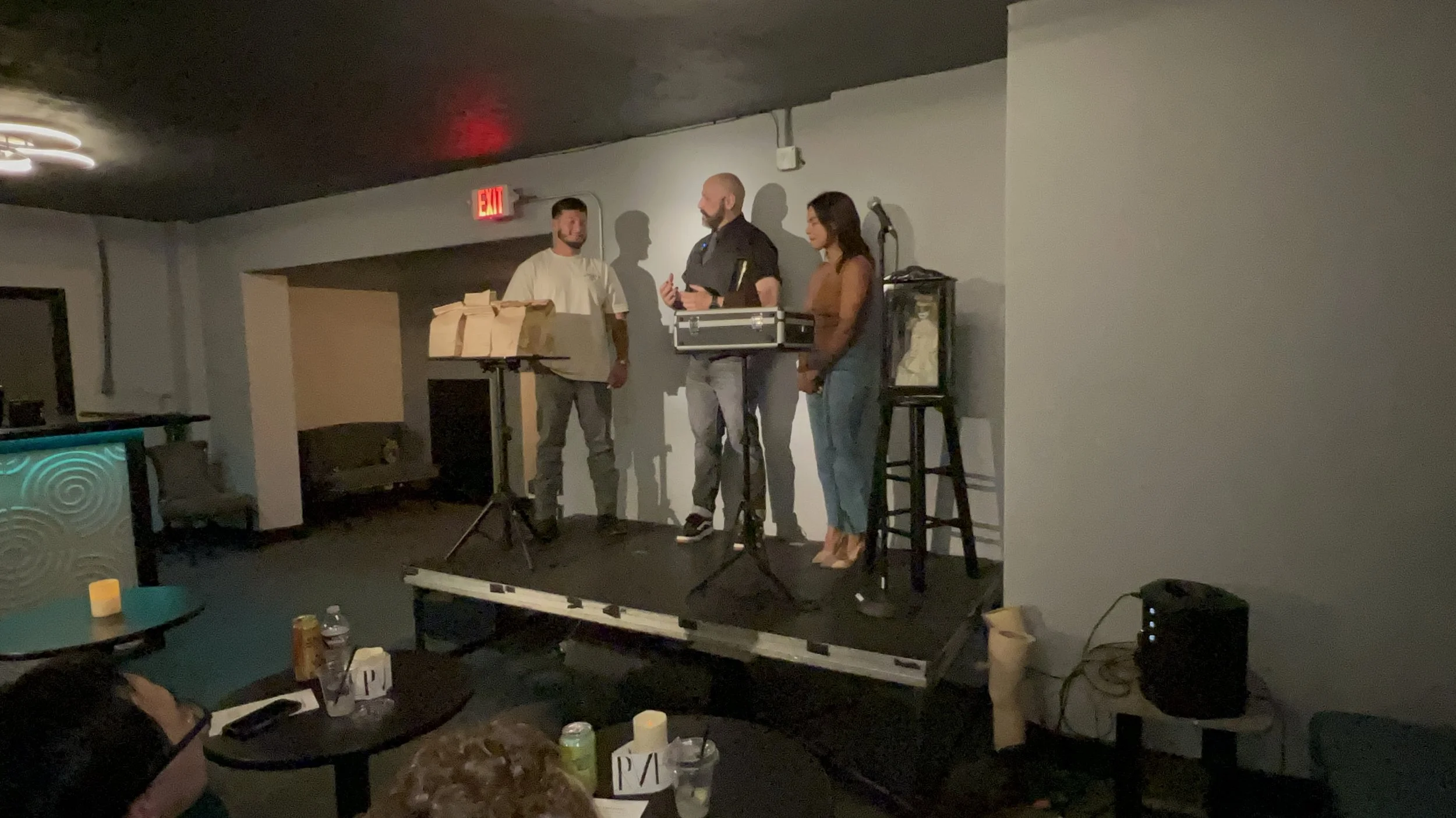 Three people on a small stage in a dimly lit room, two men and one woman. One man is in a white shirt, the other in a dark shirt and gray pants, holding a microphone. The woman is dressed in a sleeveless top and jeans. There are tables with drinks an
