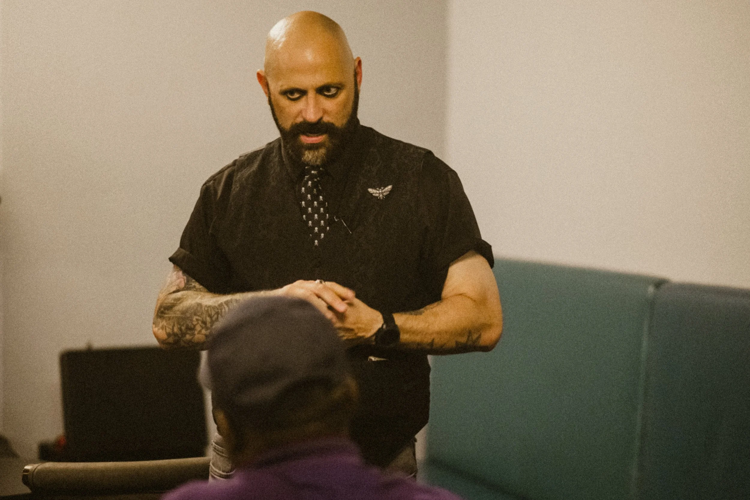 A bald man with a beard and tattoos on his arms, wearing a black shirt with a winged pin and a black watch, stands with his hands clasped in front of him, looking intently at a person seated in front of him in a room with plain walls.