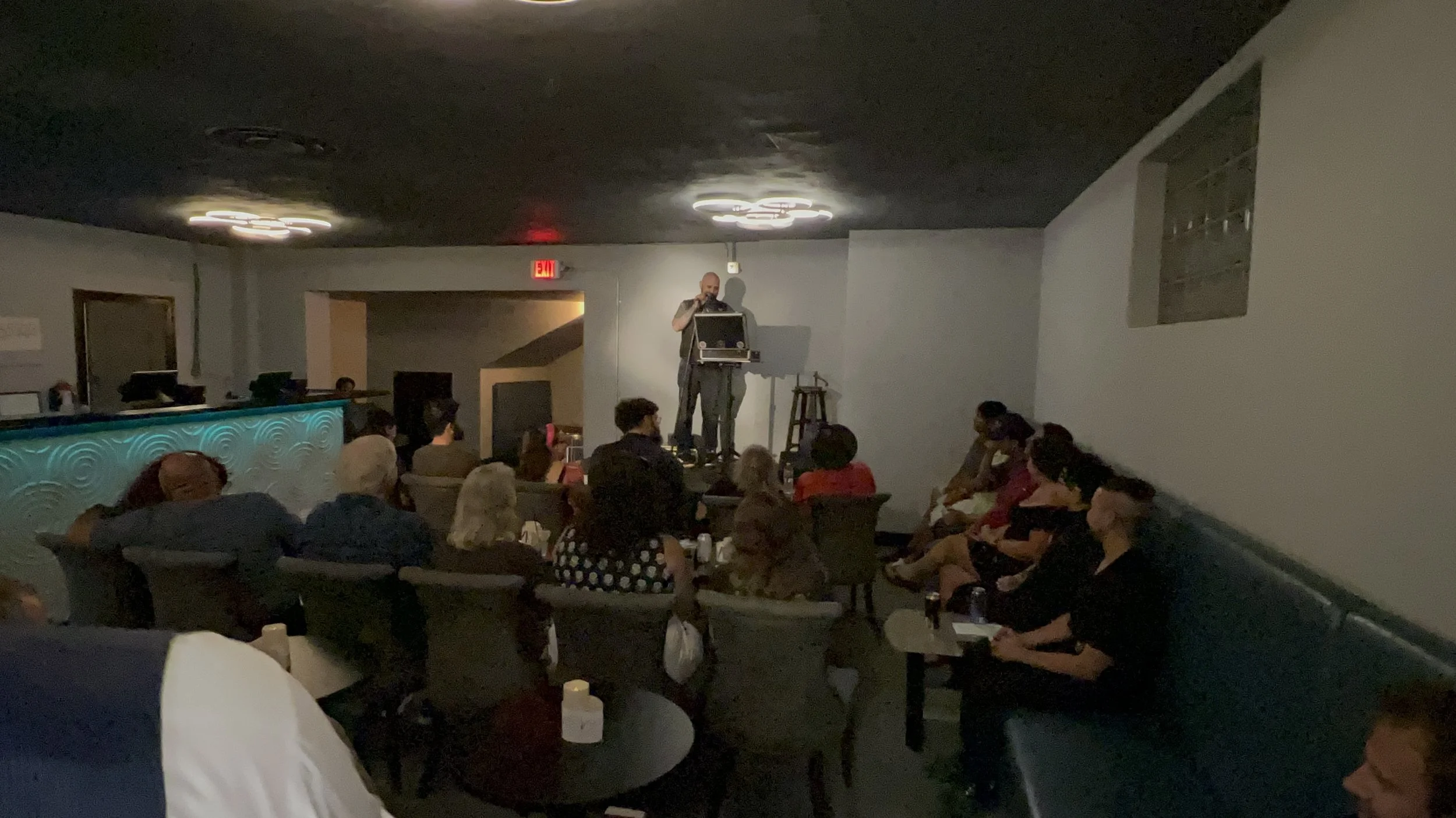A comedy show is taking place in a dimly lit venue. A performer on stage holds a microphone and uses a laptop, standing next to a stool. Audience members are seated on chairs and benches, watching the performance.