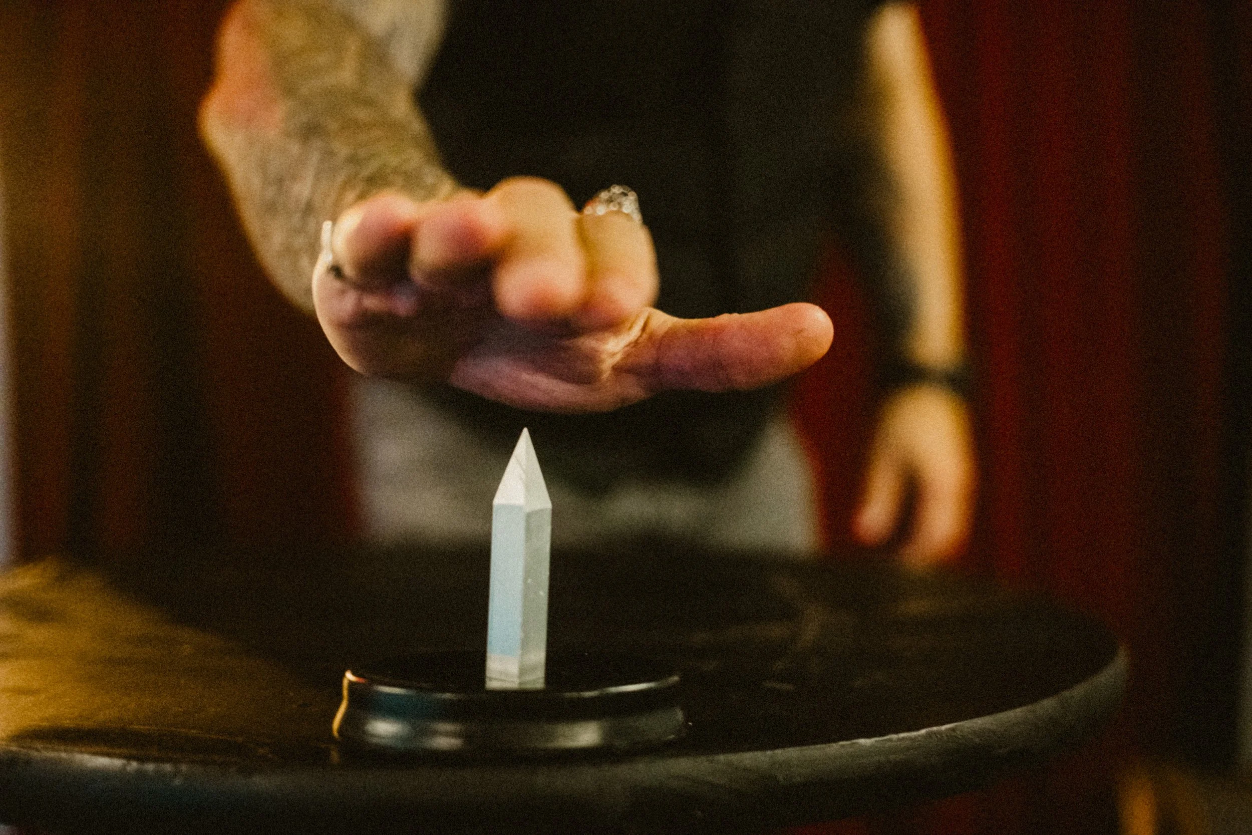 Person's hand reaching towards a pointed steel spike on a black surface, with a blurred background.
