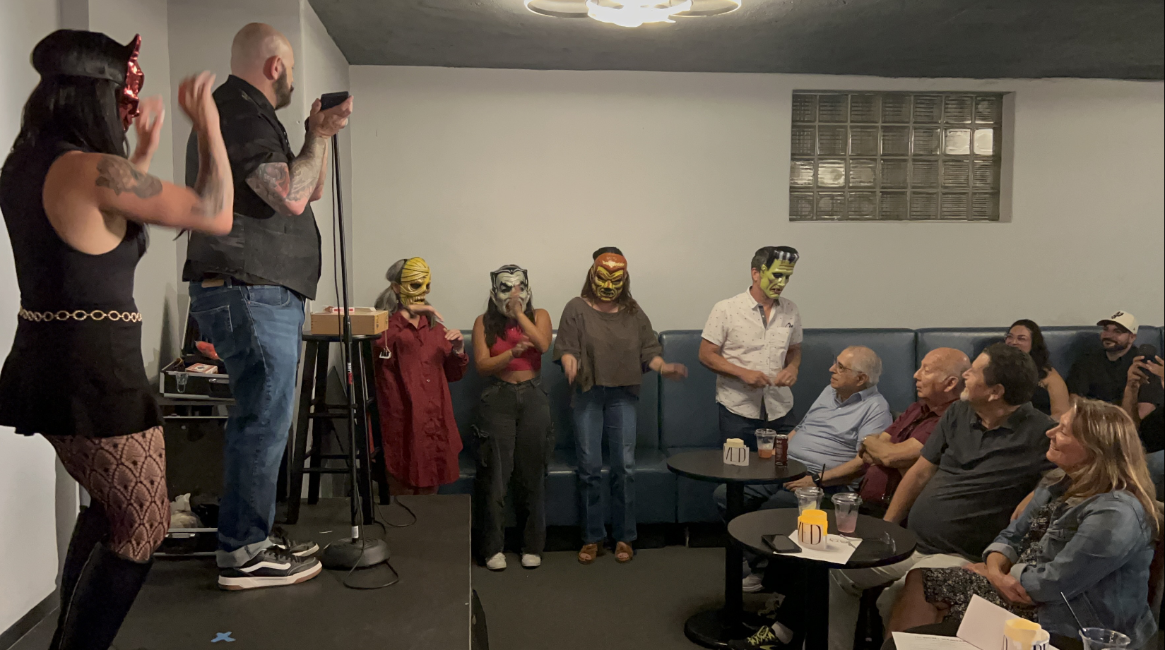 A group of five people wearing colorful masks perform on a small stage for an audience seated at tables, with some audience members taking pictures and watching the performance.