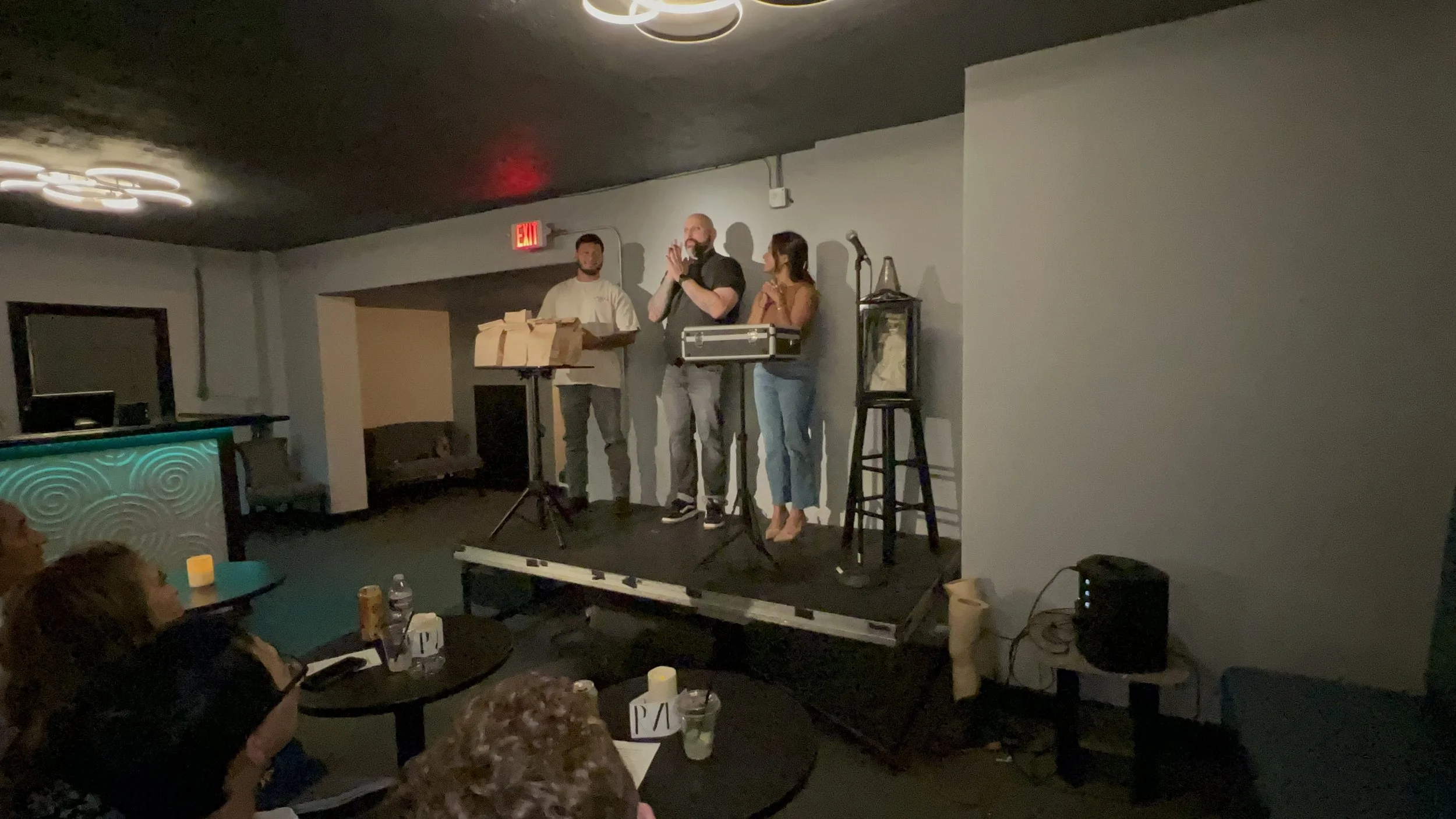 A comedy club stage with three performers standing; one man is clapping, and one woman is speaking into a microphone. There are audience members seated at tables watching, with drinks and a candle on the tables. The stage has a microphone stand and m