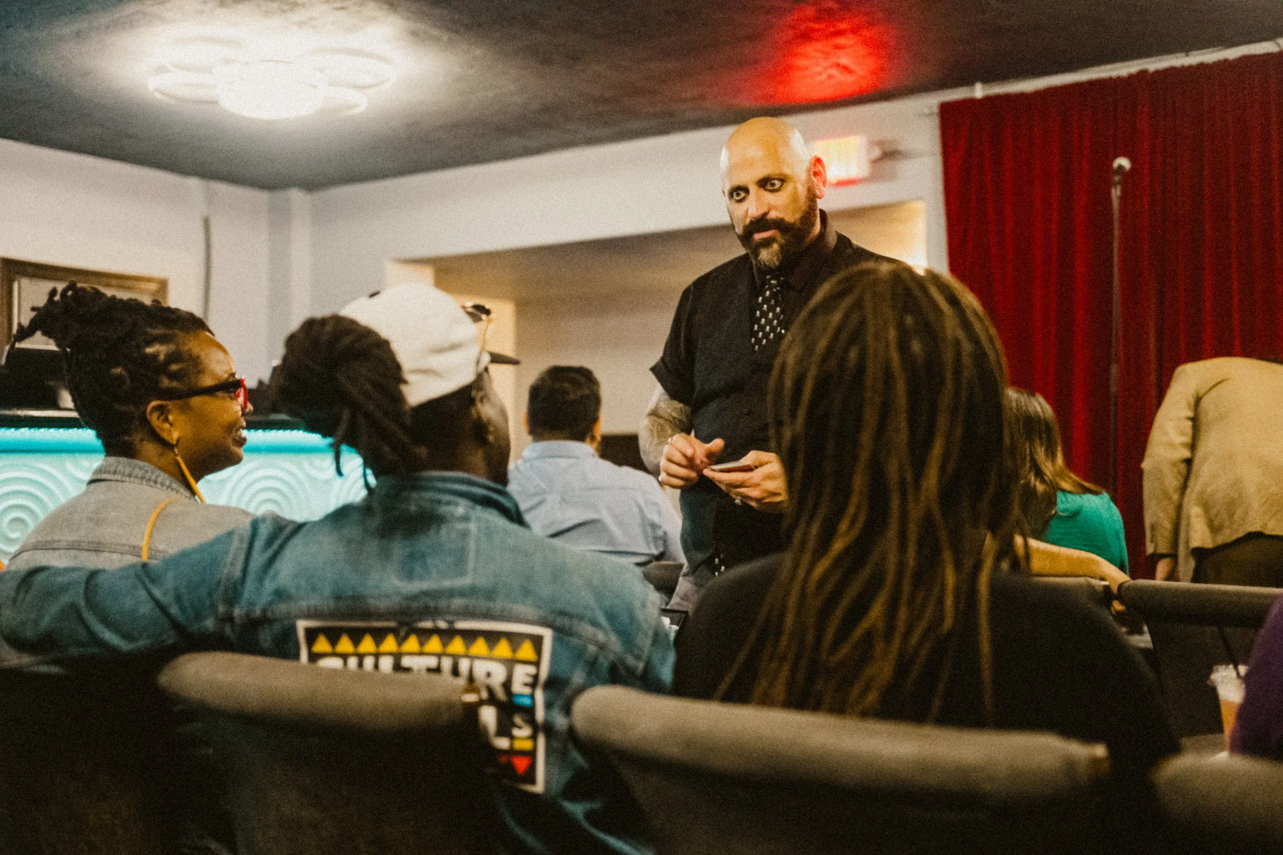 A man with a shaved head and beard wearing a black shirt with rolled sleeves and a polka dot tie, engaging in conversation with a woman with dreadlocks, seated in a room with a red curtain in the background and several other people.