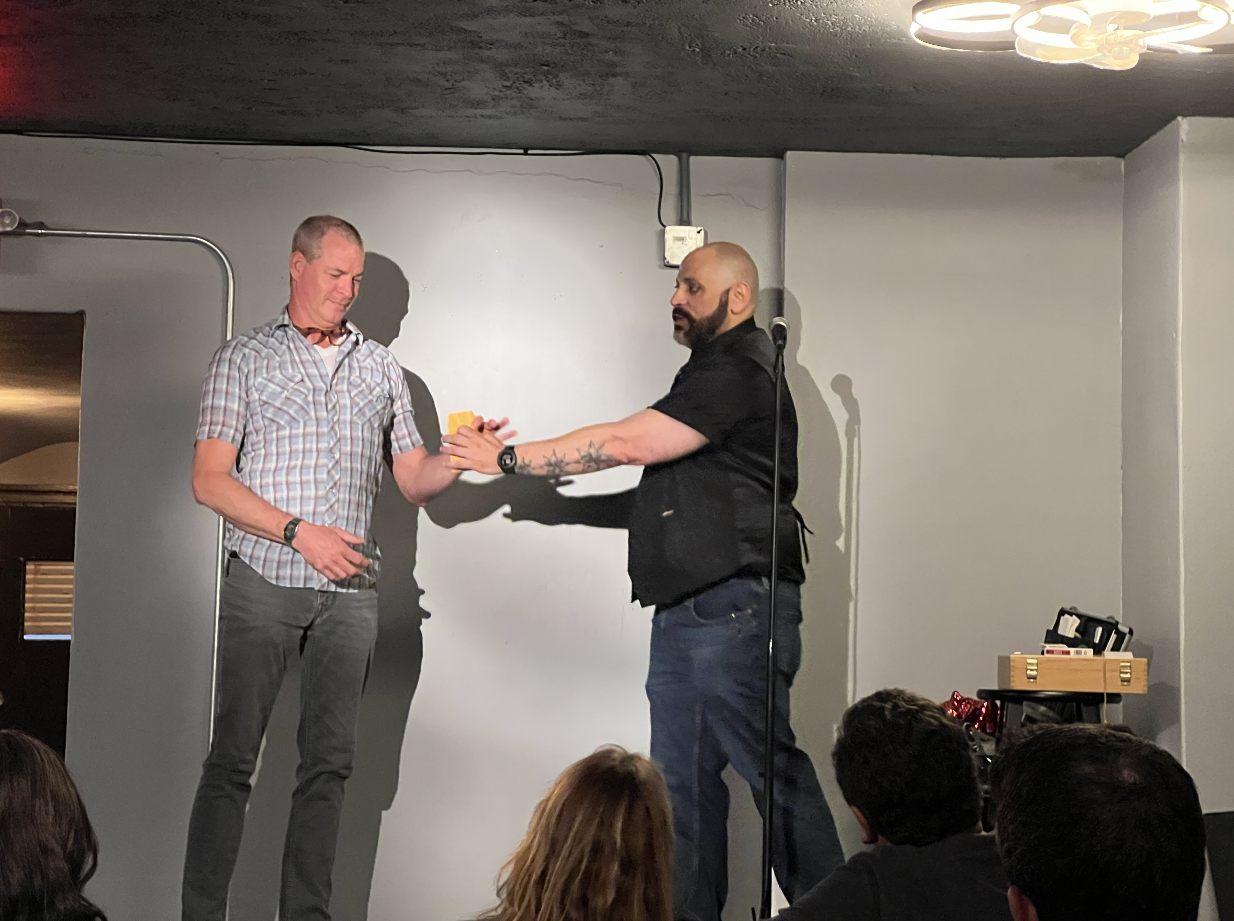 A man is handing a yellow object to another man on stage during an event or performance, with an audience watching.