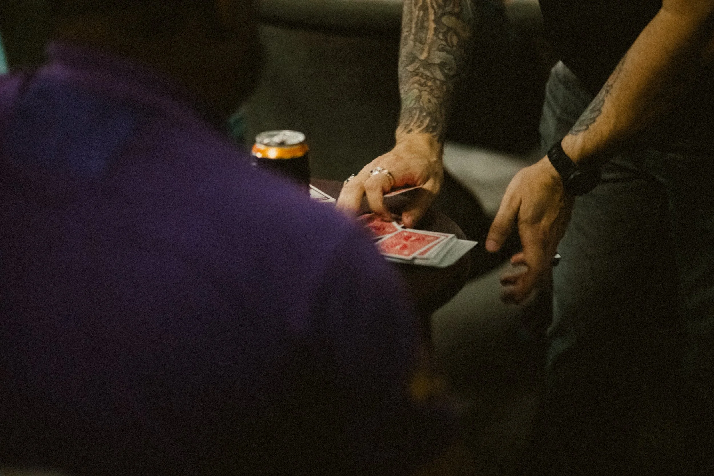 People playing cards at a table, with one person's arm and tattoos visible, holding cards. A can of drink is on the table.