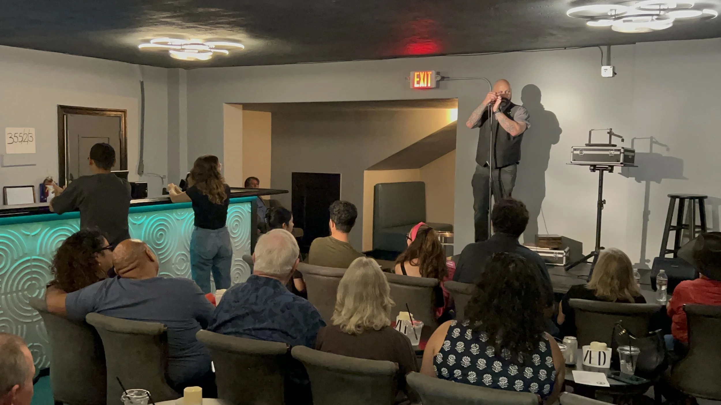 A comedian performs on stage at an indoor comedy show while an audience watches. The stage has a microphone and audio equipment, with some people in the crowd seated and drinking drinks.