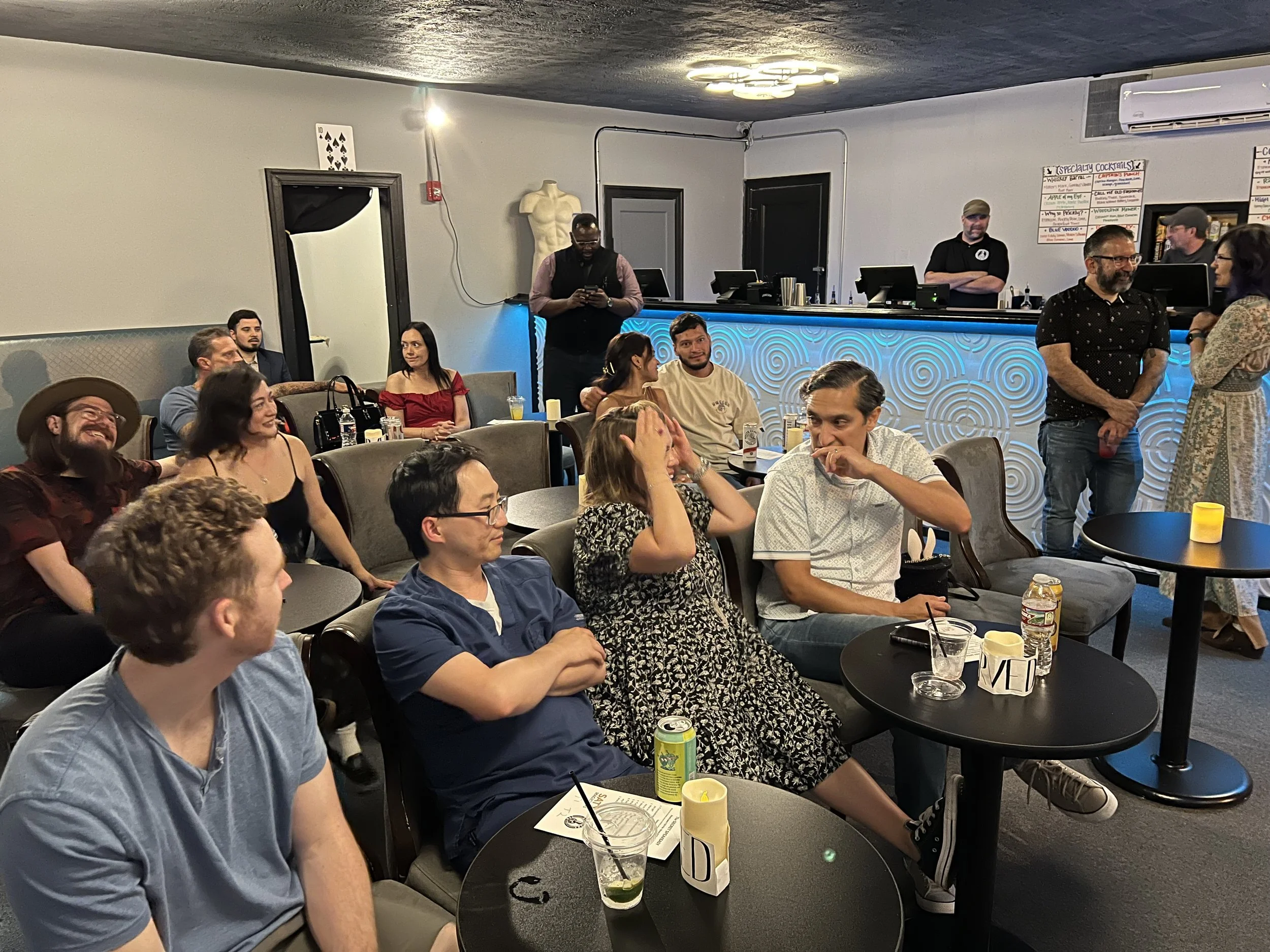 A group of people sitting at tables in a bar or lounge, some smiling and talking, with a bar counter and bartender in the background. The room has dim lighting and a casual atmosphere.