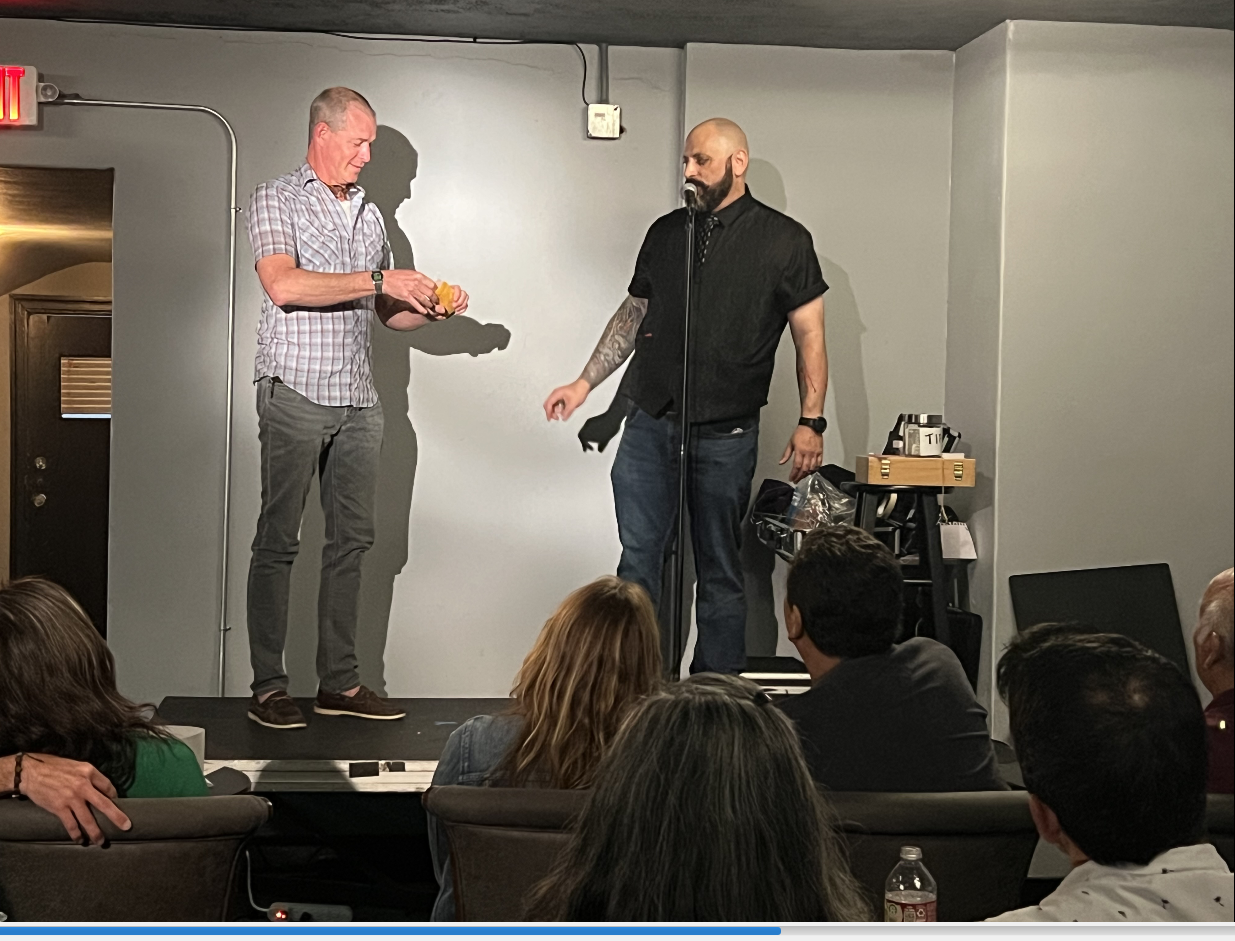 Two men stand on a small stage, one handing an object to the other, in front of an audience seated in a dimly lit room with gray walls.